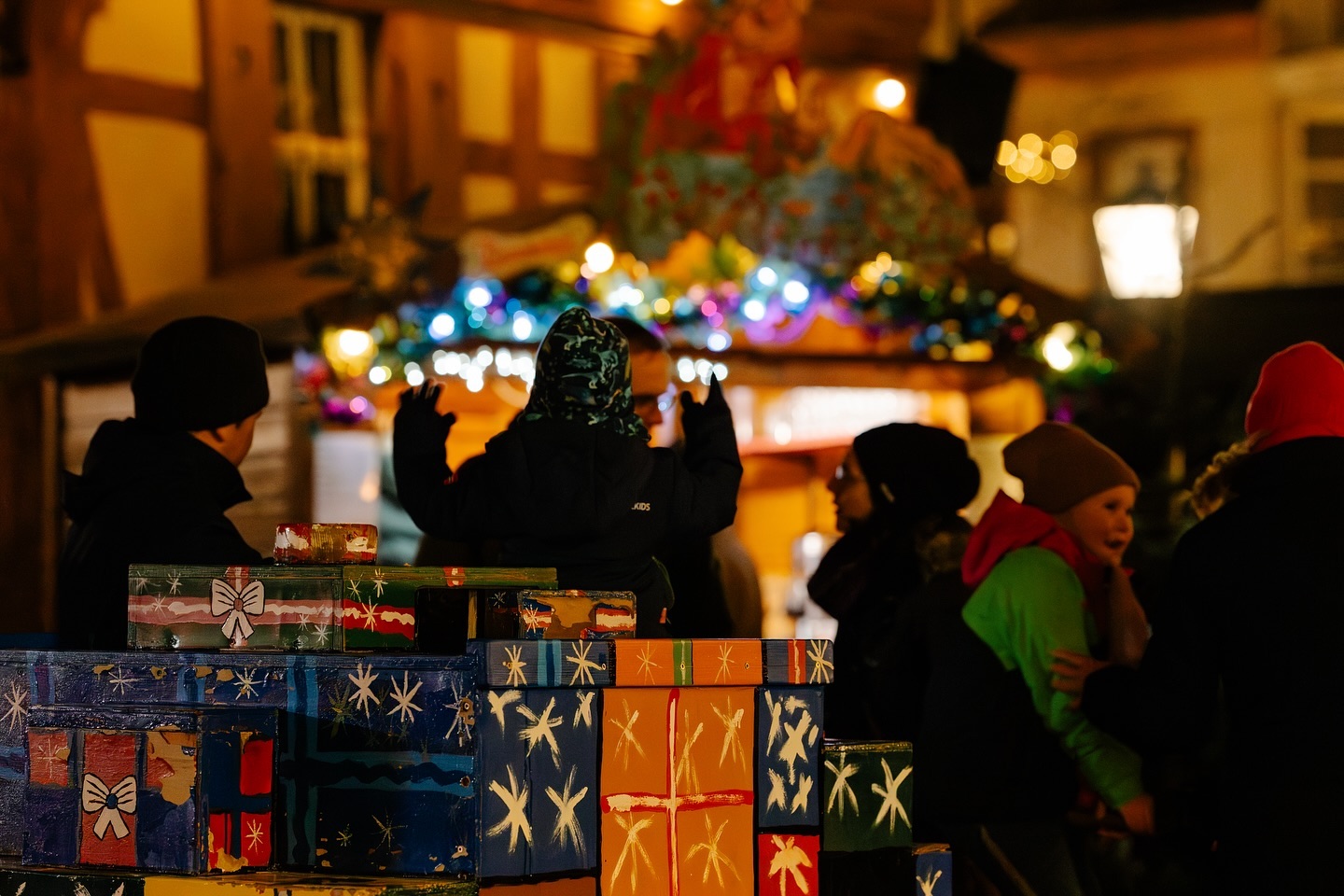 Kinder stehen vor einem Weihnachtsmarktstand mit vielen bunten Geschenkboxen.