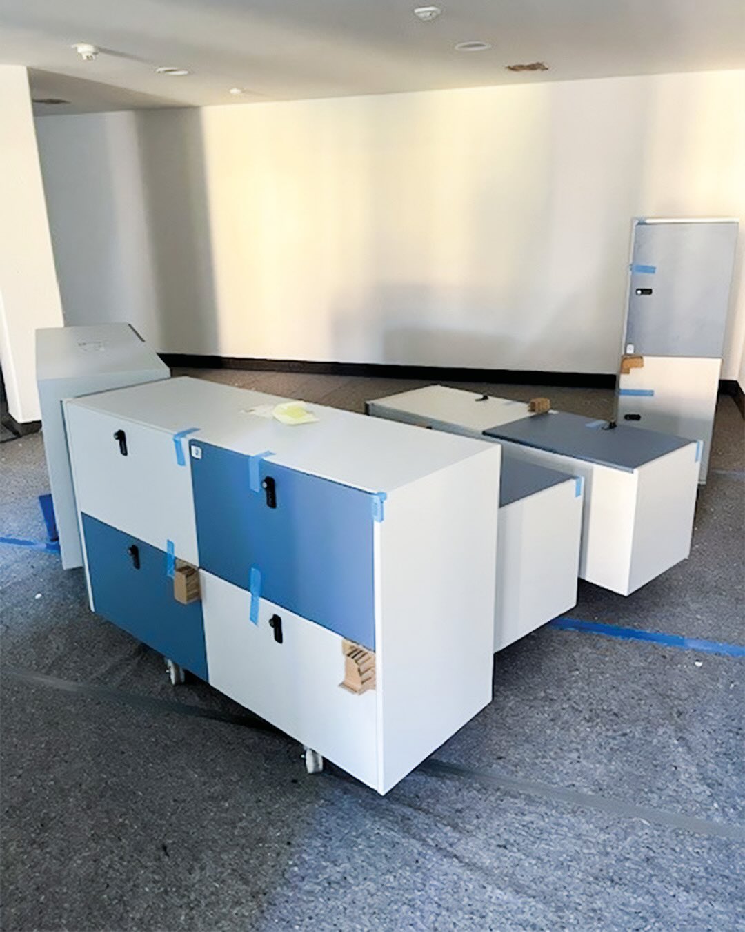 Dieser Alternativtext wurde KI-generiert: Ein Innenraum mit mehreren Schrankelementen in verschiedenen Farben. Die Elemente stehen auf Rollen und sind teilweise mit Klebeband gesichert.