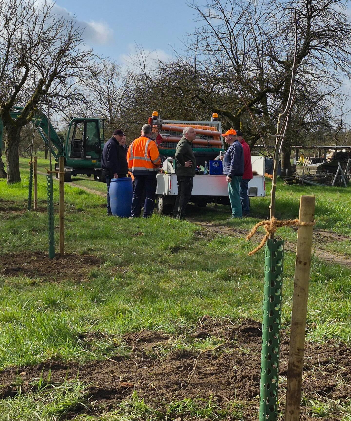 Dieser Alternativtext wurde KI-generiert: Eine Gruppe von Menschen in Arbeitskleidung steht auf einer Wiese um ein Fahrzeug und Geräte herum. Im Hintergrund ist eine Baumaschine zu sehen.