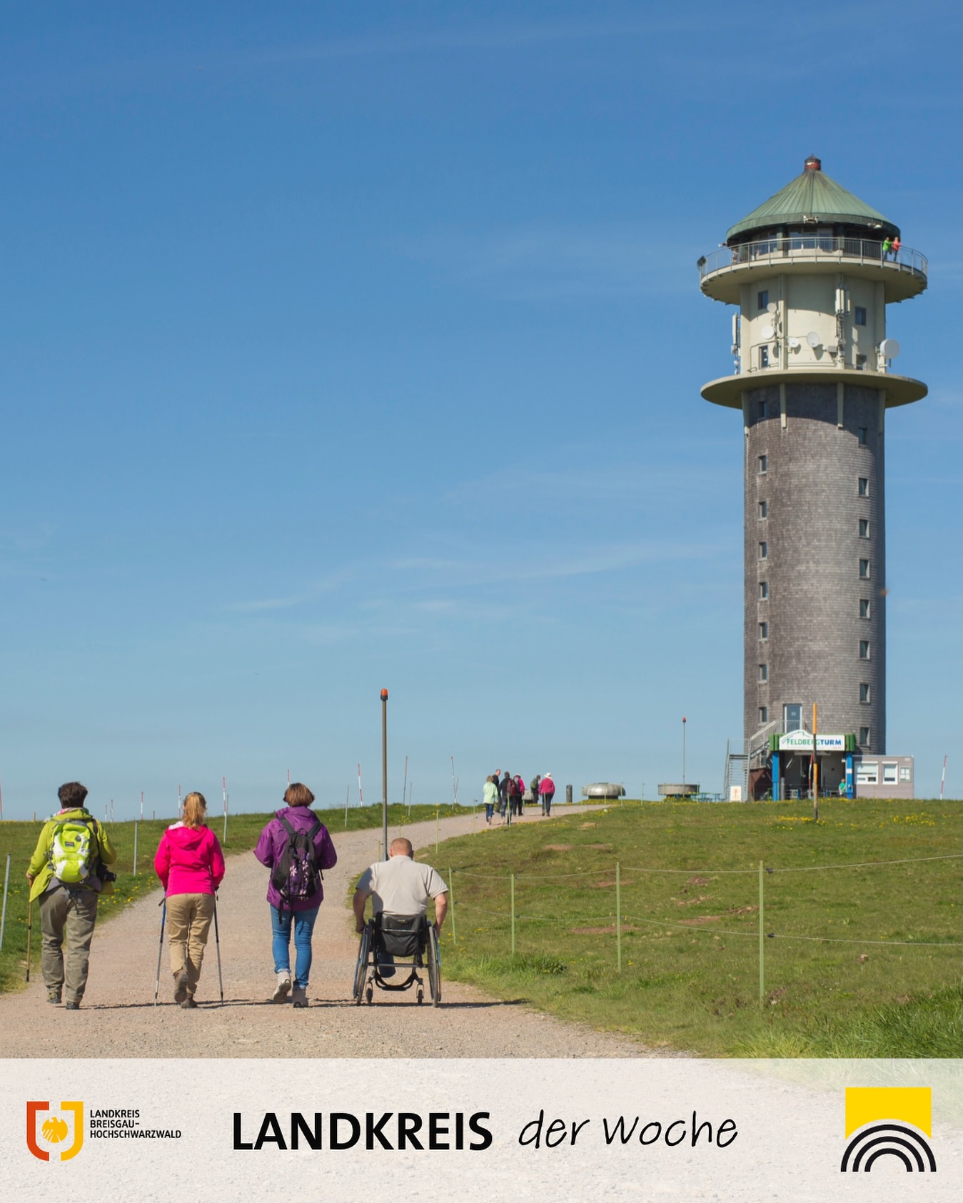 Dieser Alternativtext wurde KI-generiert: Ein hoher, grauer Turm steht auf einer grünen Hügellandschaft. Mehrere Personen sind auf einem Weg zu sehen, einige gehen, eine Person sitzt in einem Rollstuhl.