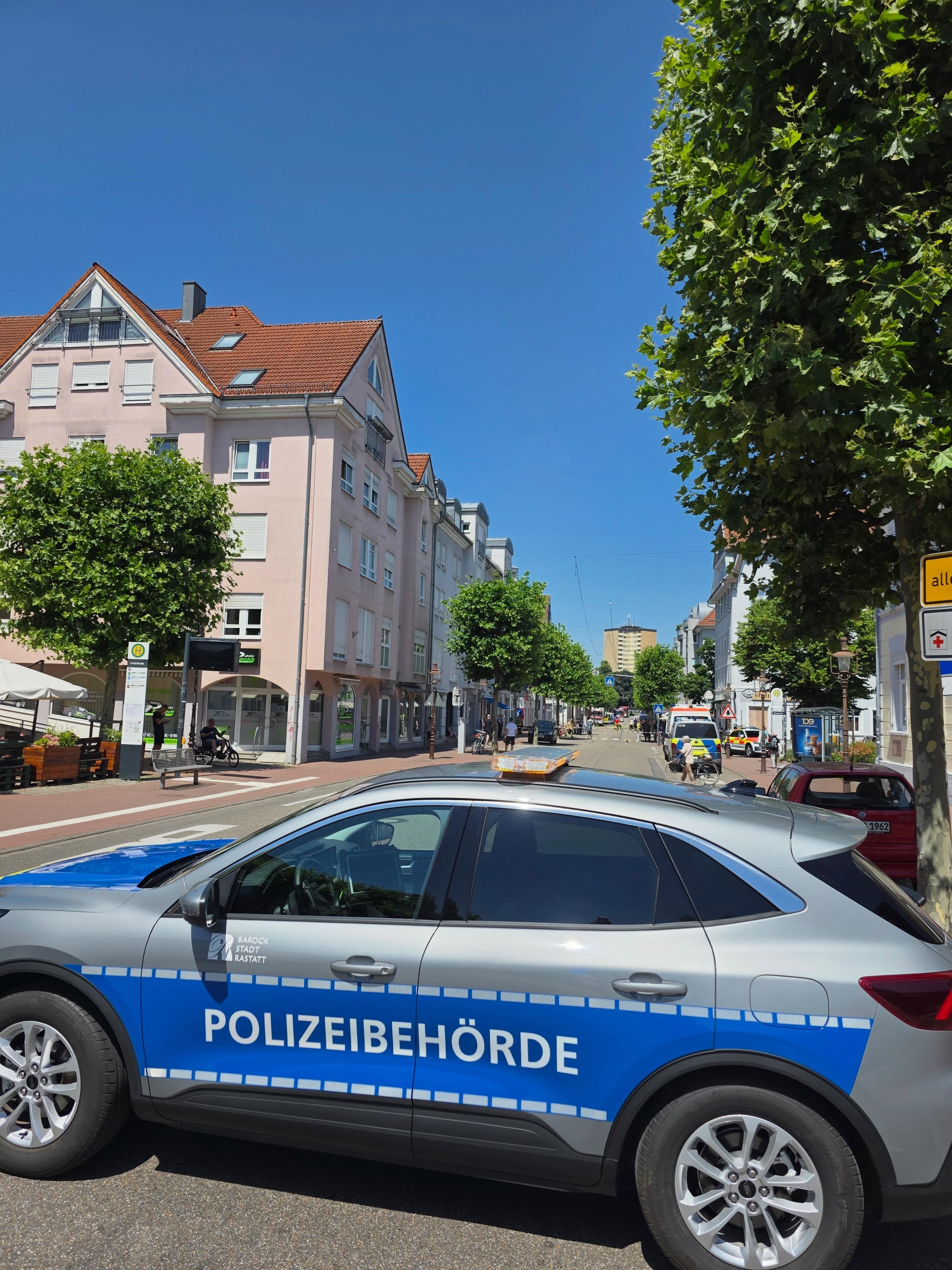 Ein silberner Polizeiwagen steht im Vordergrund auf einer Straße, auf dessen blauer Seitenleiste "POLIZEIBEHÖRDE" und oben "BAROCK STADT RASTATT" steht. Der Hintergrund zeigt eine sonnige Straßenszene mit rosa Mehrfamilienhäusern, Bäumen und weiteren Fahrzeugen, darunter einen Rettungswagen in der Ferne. (KI-Alternativtext)