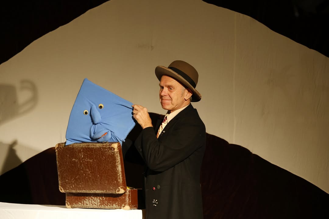 Dieser Alternativtext wurde KI-generiert: Eine Person mit Hut hält eine blaue Stofffigur vor einer Kiste. Im Hintergrund ist ein Schattenwurf zu sehen.