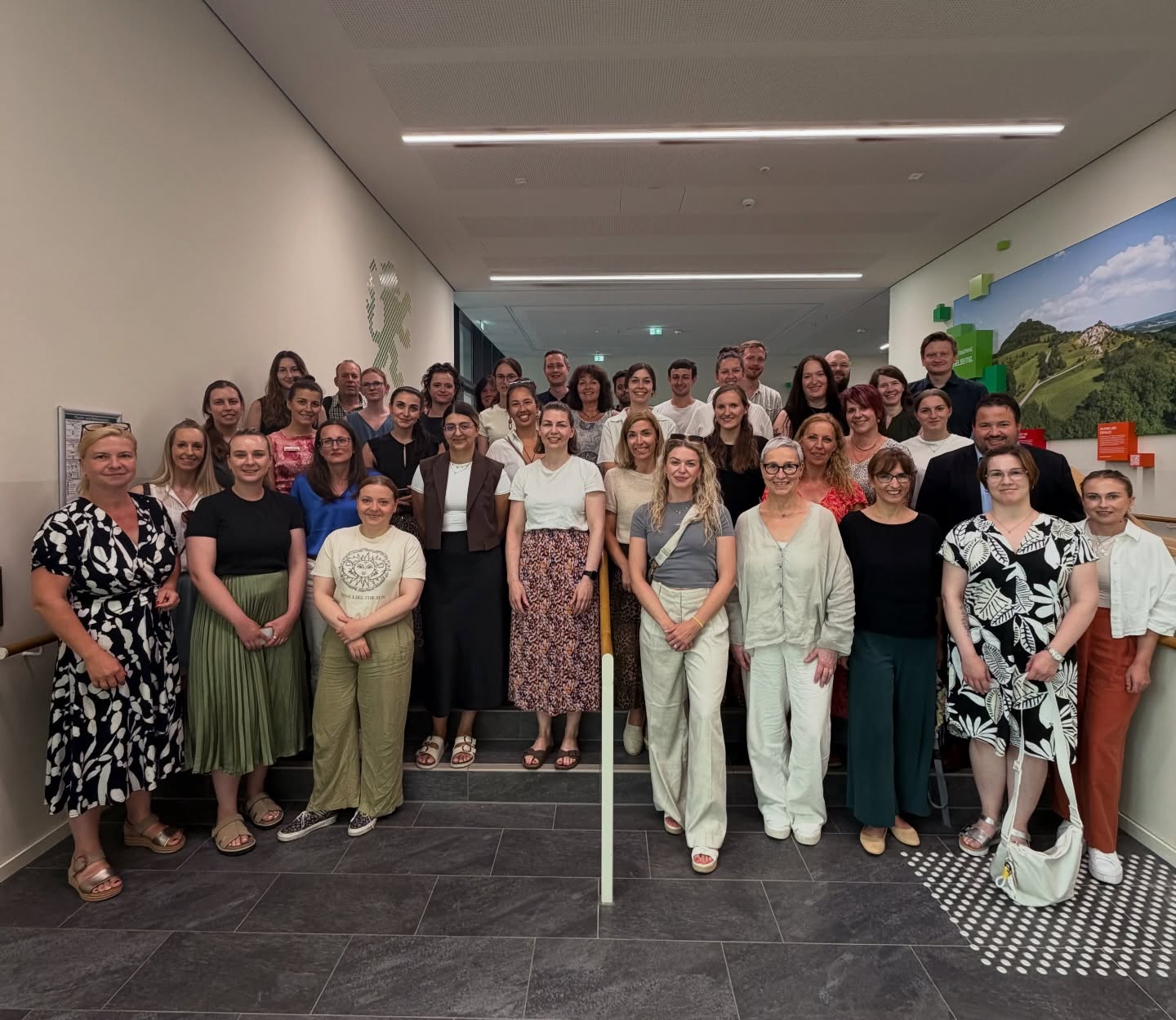 Eine große Gruppe von Menschen steht in mehreren Reihen auf einer Treppe und im Flur und posiert lächelnd für ein Gruppenfoto. An der rechten Wand hängt ein großes Bild einer grünen Landschaft. (KI-Alternativtext)