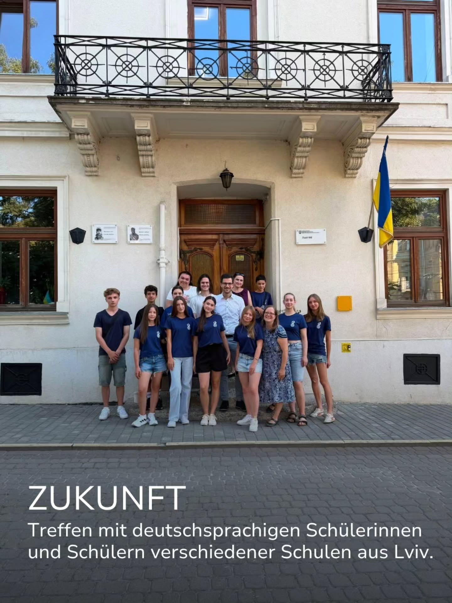 Eine Gruppe von Personen, die größtenteils blaue T-Shirts tragen, posiert vor einem Gebäude mit ukrainischer Flagge. Im unteren Bildbereich steht der Text: „ZUKUNFT Treffen mit deutschsprachigen Schülerinnen und Schülern verschiedener Schulen aus Lviv.“ (KI-Alternativtext)