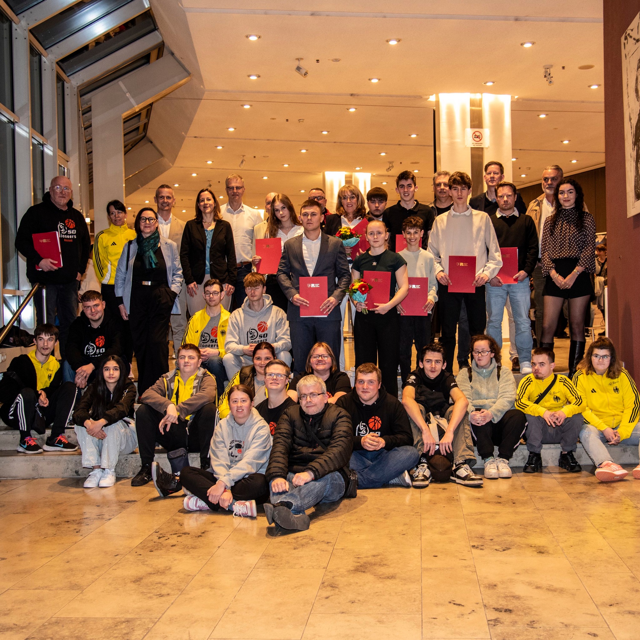 Dieser Alternativtext wurde KI-generiert: Eine große Gruppe von Menschen steht auf einer hellen, gefliesten Fläche vor einer roten Wand und einer Treppe. Einige halten Dokumente oder Blumen in den Händen.
