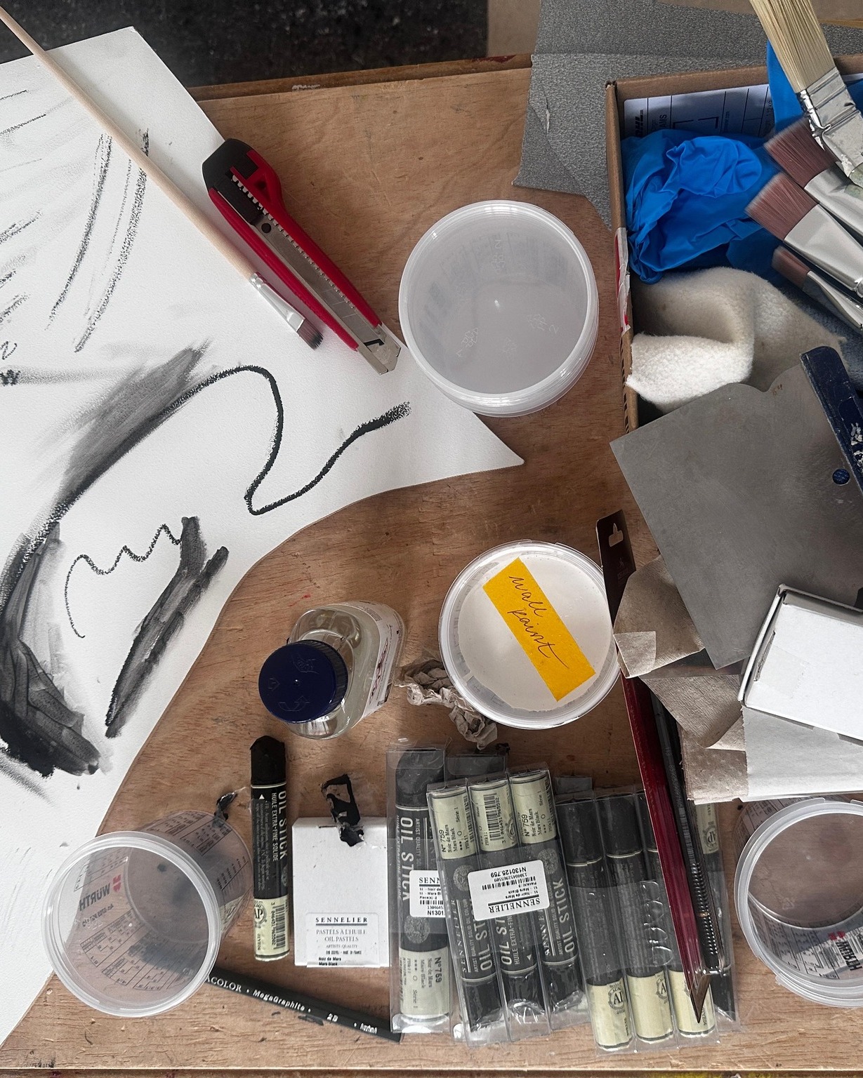 Dieser Alternativtext wurde KI-generiert: Schwarz-Weiß-Foto einer Arbeitsfläche mit verschiedenen Kunstmaterialien, darunter Stifte, Farben und Behälter. Die Gegenstände sind unordentlich angeordnet und lassen auf kreative Arbeit schließen.