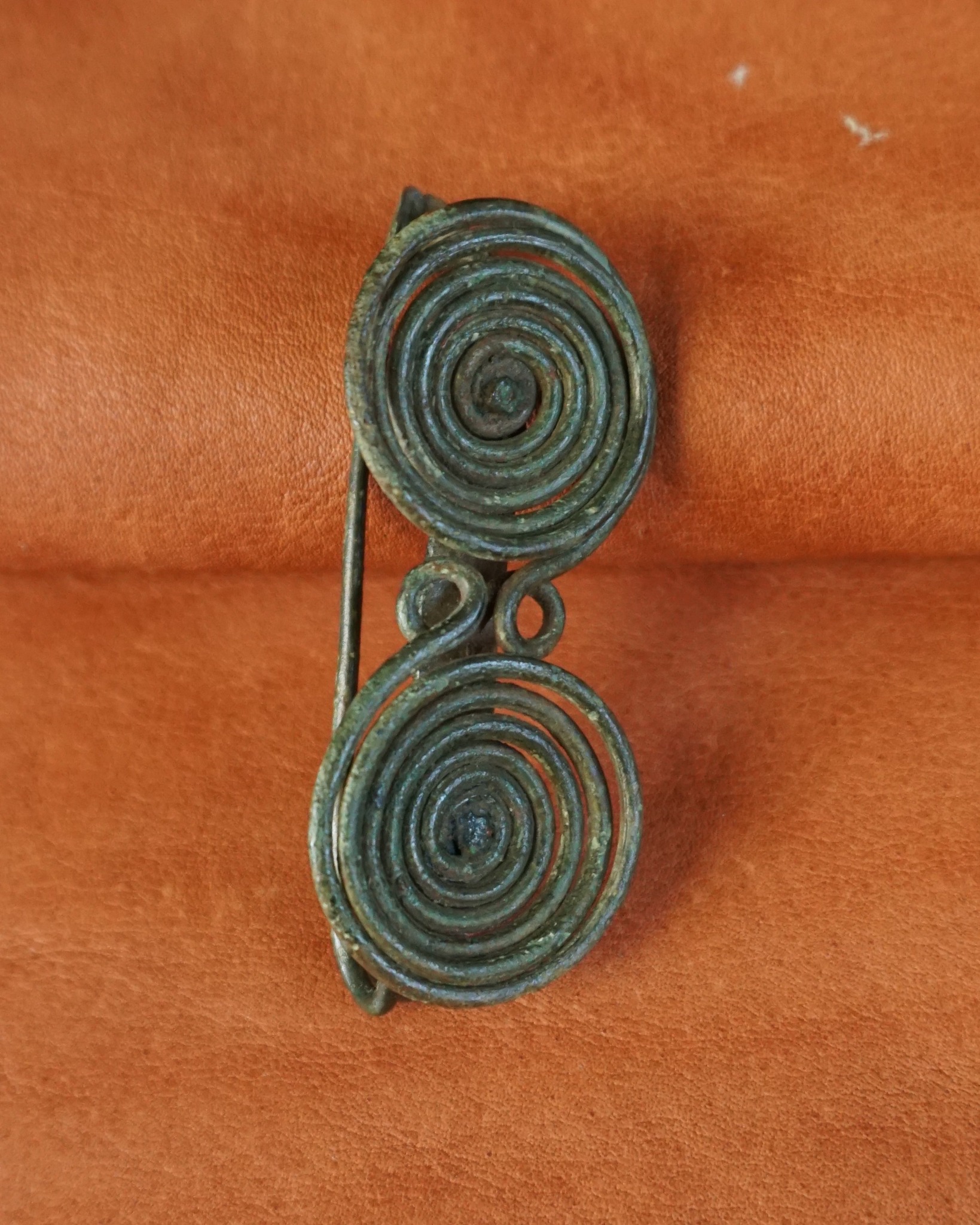 Eine bronzene Brosche mit zwei großen Spiralen und grüner Patina liegt auf einem orangefarbenen Hintergrund. Die Spiralen sind durch ein Drahtstück mit kleinen Schlaufen verbunden, und eine Nadel ist an der linken Seite sichtbar. (KI-Alternativtext)