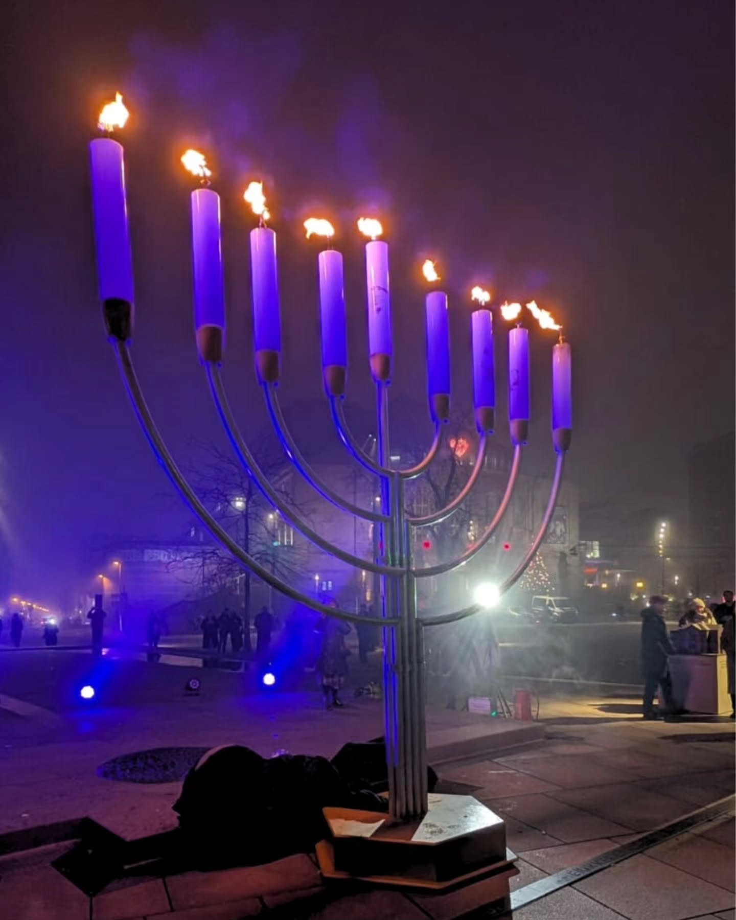 Dieser Alternativtext wurde KI-generiert: Eine Ansammlung von leuchtenden, lilafarbenen Kerzen steht auf einem silberfarbenen Gestell. Im Hintergrund sind verschwommene Lichter und Menschen zu erkennen.