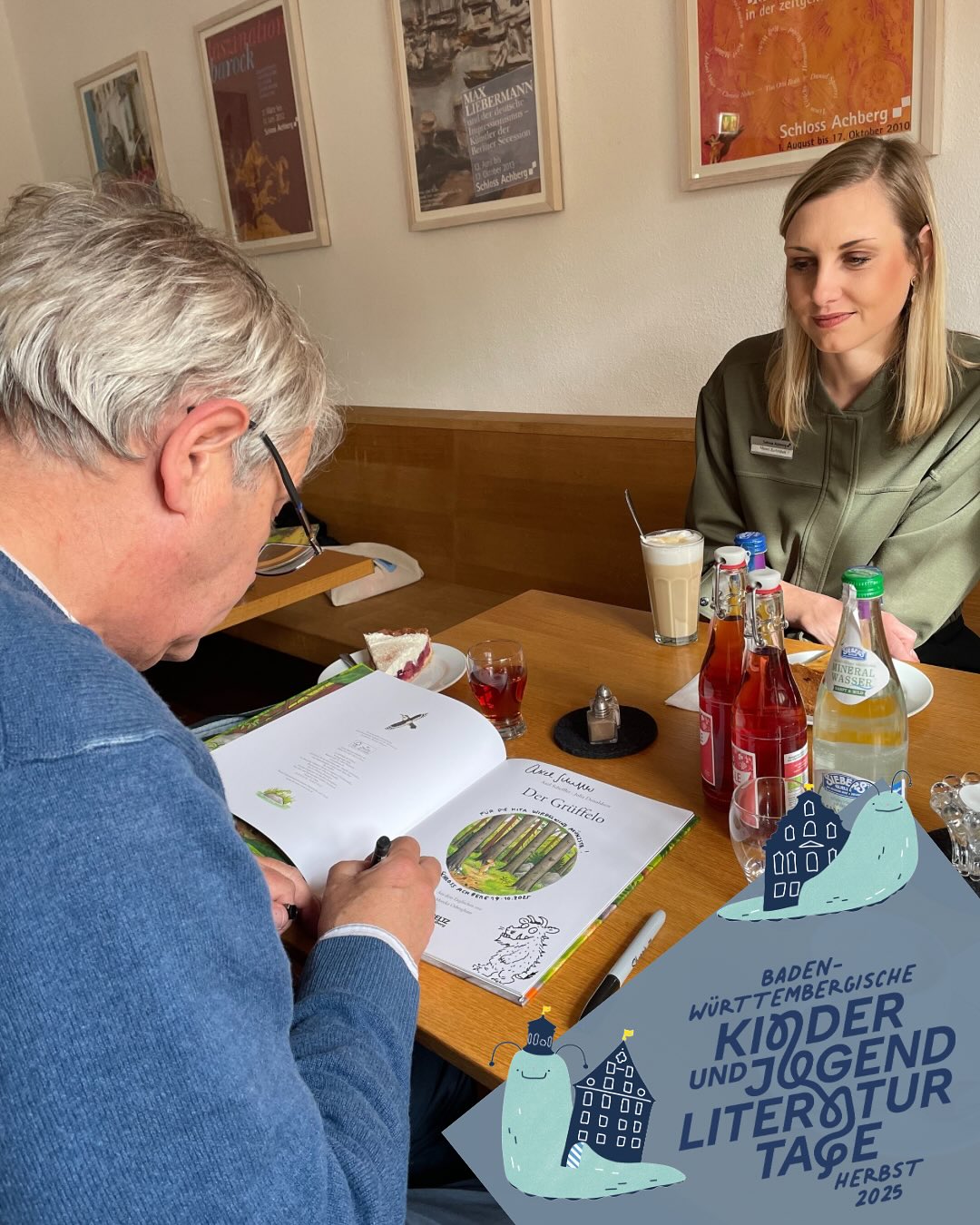 Dieser Alternativtext wurde KI-generiert: Zwei Personen sitzen an einem Tisch in einem hellen Raum und unterhalten sich. Auf dem Tisch befinden sich Getränke, Essen und ein aufgeschlagenes Buch mit Illustrationen.