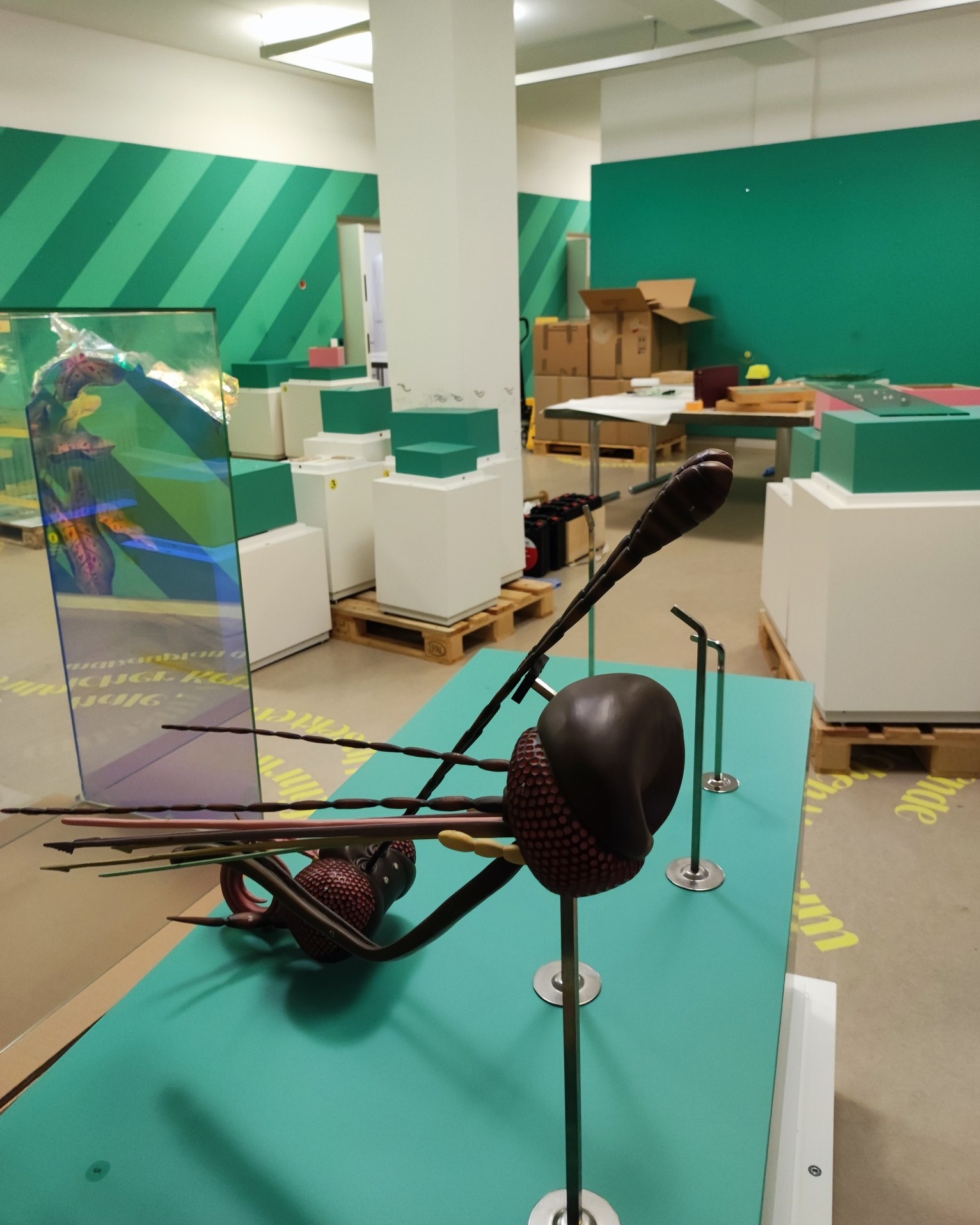 Dieser Alternativtext wurde KI-generiert: Ein Innenraum mit grünen Akzenten und einer Installation im Vordergrund. Zu sehen ist eine skulpturale Darstellung eines Insekts auf einem Sockel.