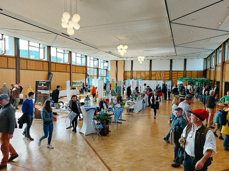 Ein großes Versammlungsgebäude, in dem viele Menschen an verschiedenen Ständen stehen und sich unterhalten. Der Boden ist aus Holz.