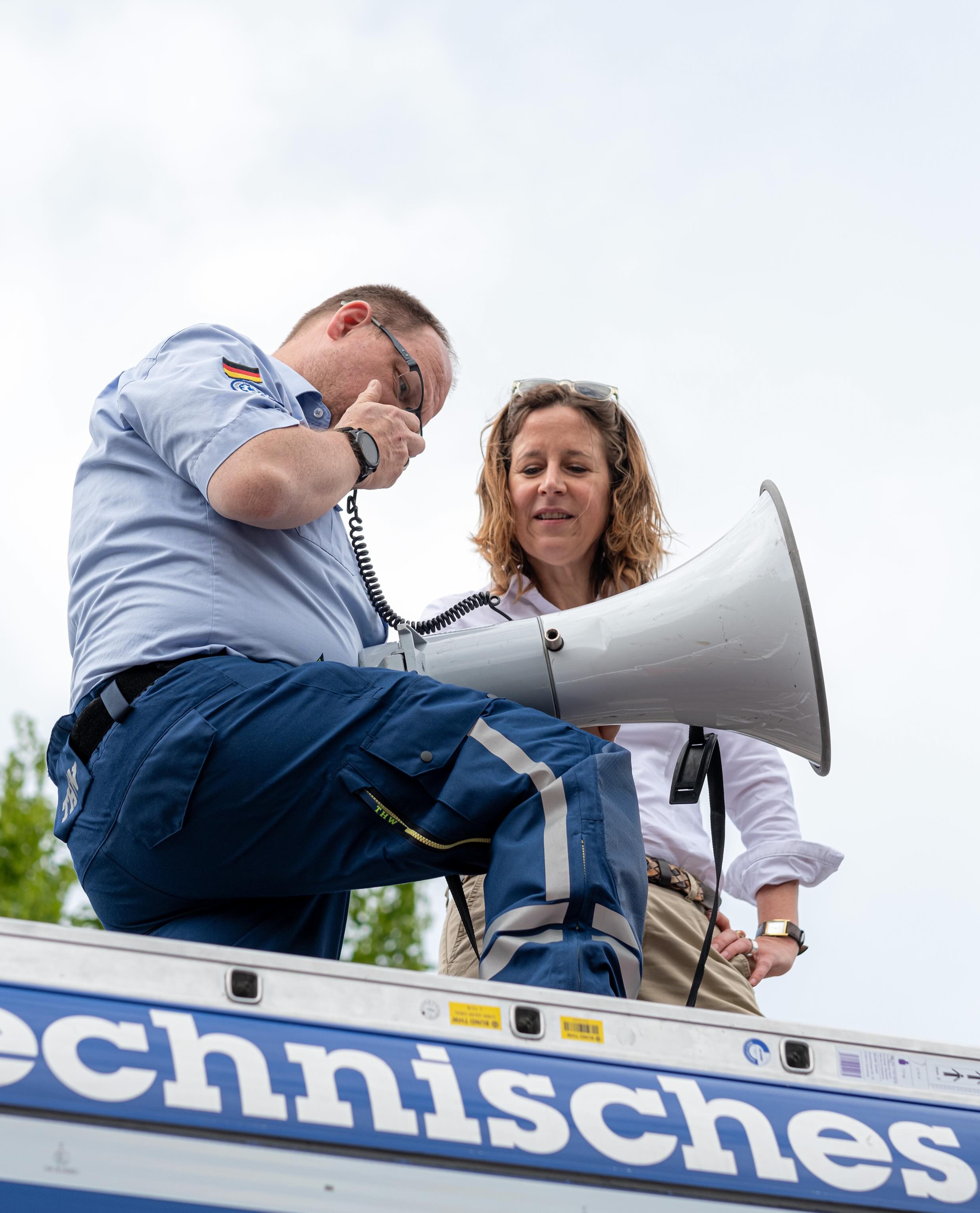 Dieser Alternativtext wurde KI-generiert: Zwei Personen in Arbeitskleidung stehen auf einem Fahrzeug. Eine Person hält ein Funkgerät, die andere ein Megafon.