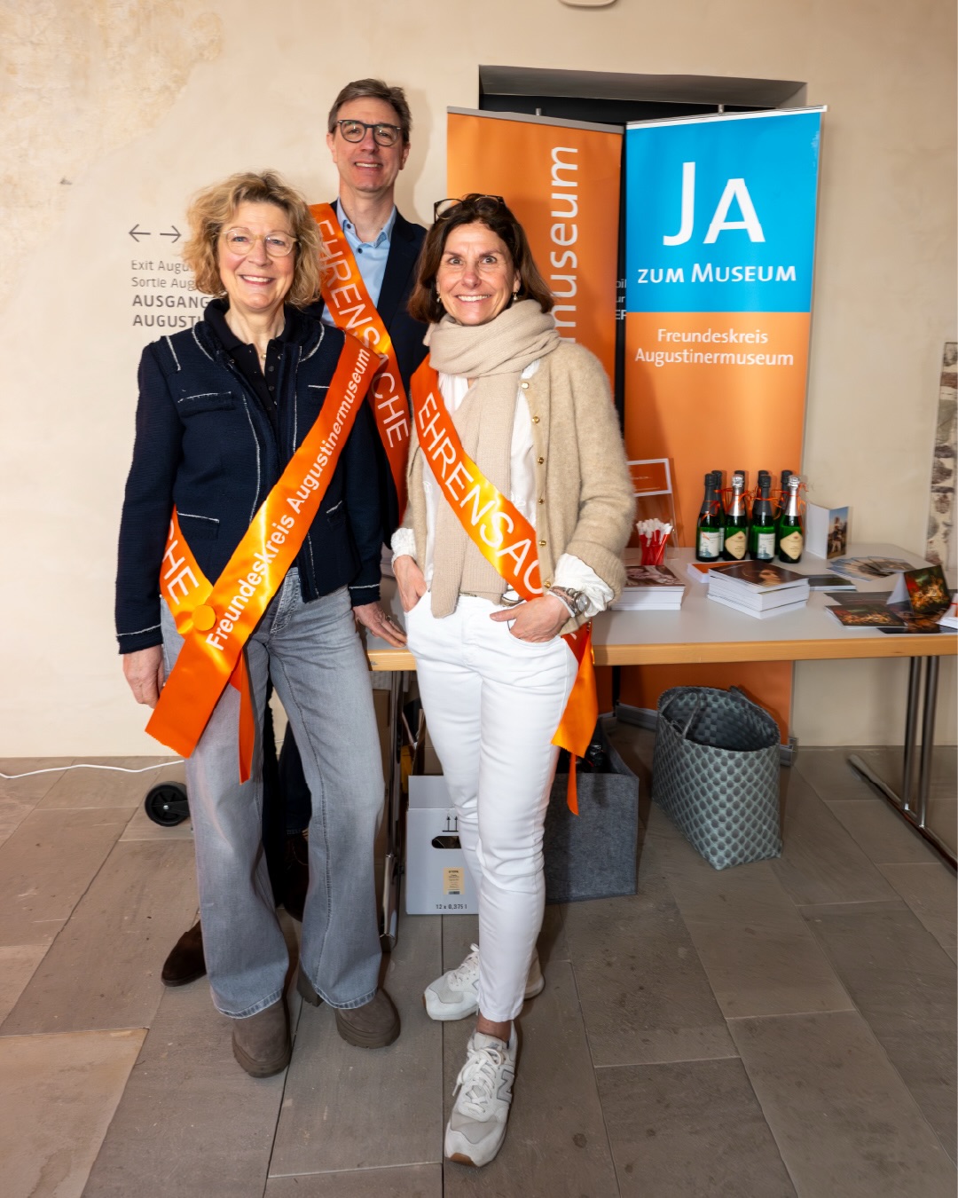 Dieser Alternativtext wurde KI-generiert: Drei Personen stehen nebeneinander vor einer Tür und einem Tisch mit Gegenständen darauf. Sie tragen orangefarbene Bänder und stehen vor einem Museumseingang.