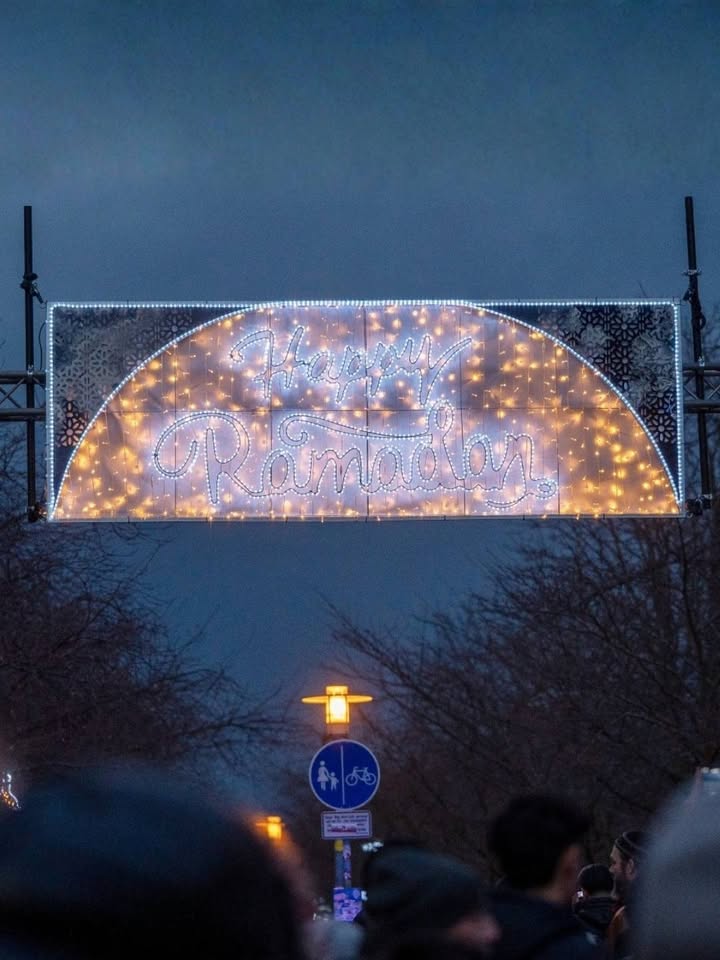 Dieser Alternativtext wurde KI-generiert: Ein beleuchteter Bogen mit der Aufschrift "Happy Ramadan". Die Aufnahme ist im Zwielicht und zeigt schemenhaft Menschen im Vordergrund.
