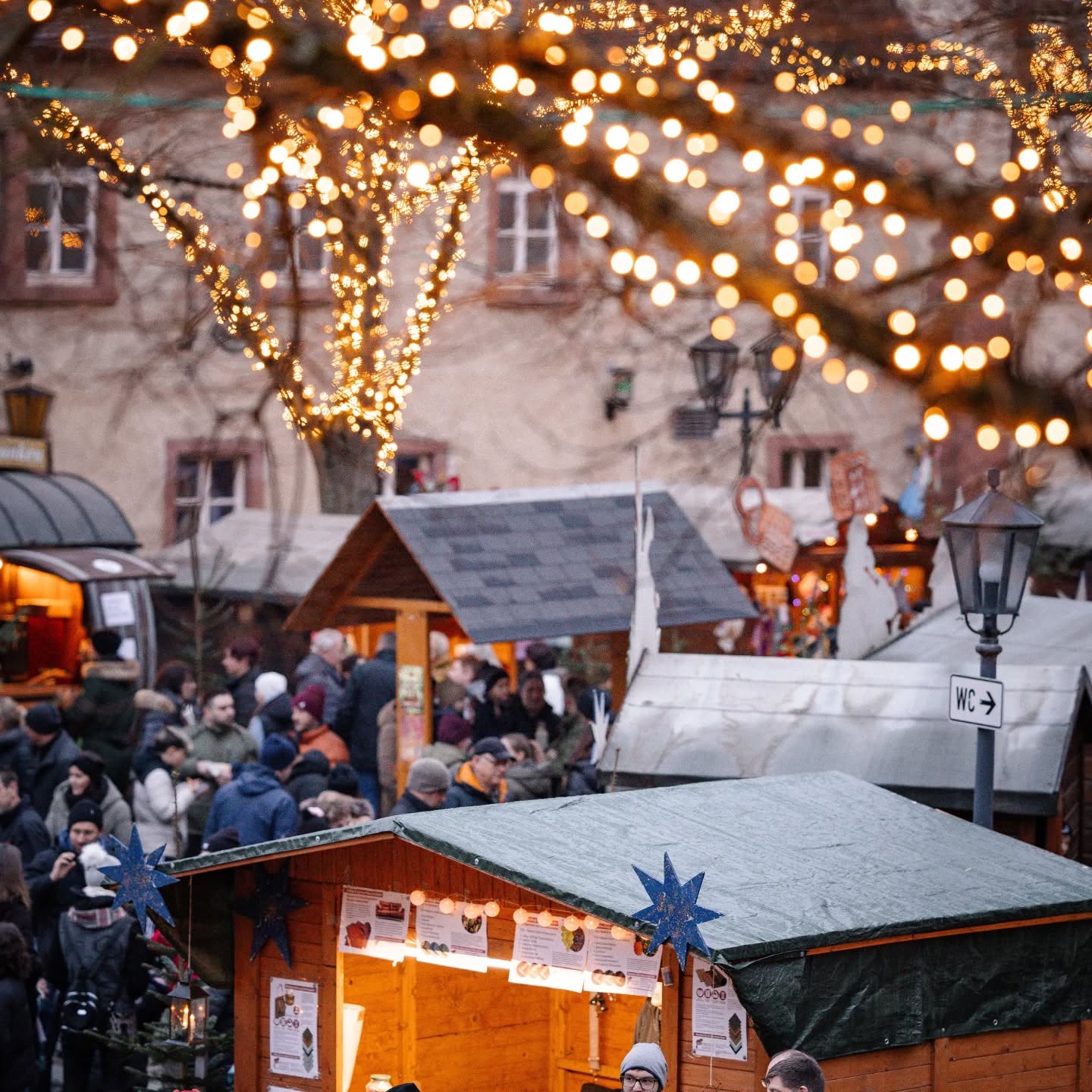 Dieser Alternativtext wurde KI-generiert: Ein belebter Marktplatz mit kleinen Holzhütten und festlicher Beleuchtung. Viele Menschen sind zwischen den Ständen unterwegs.