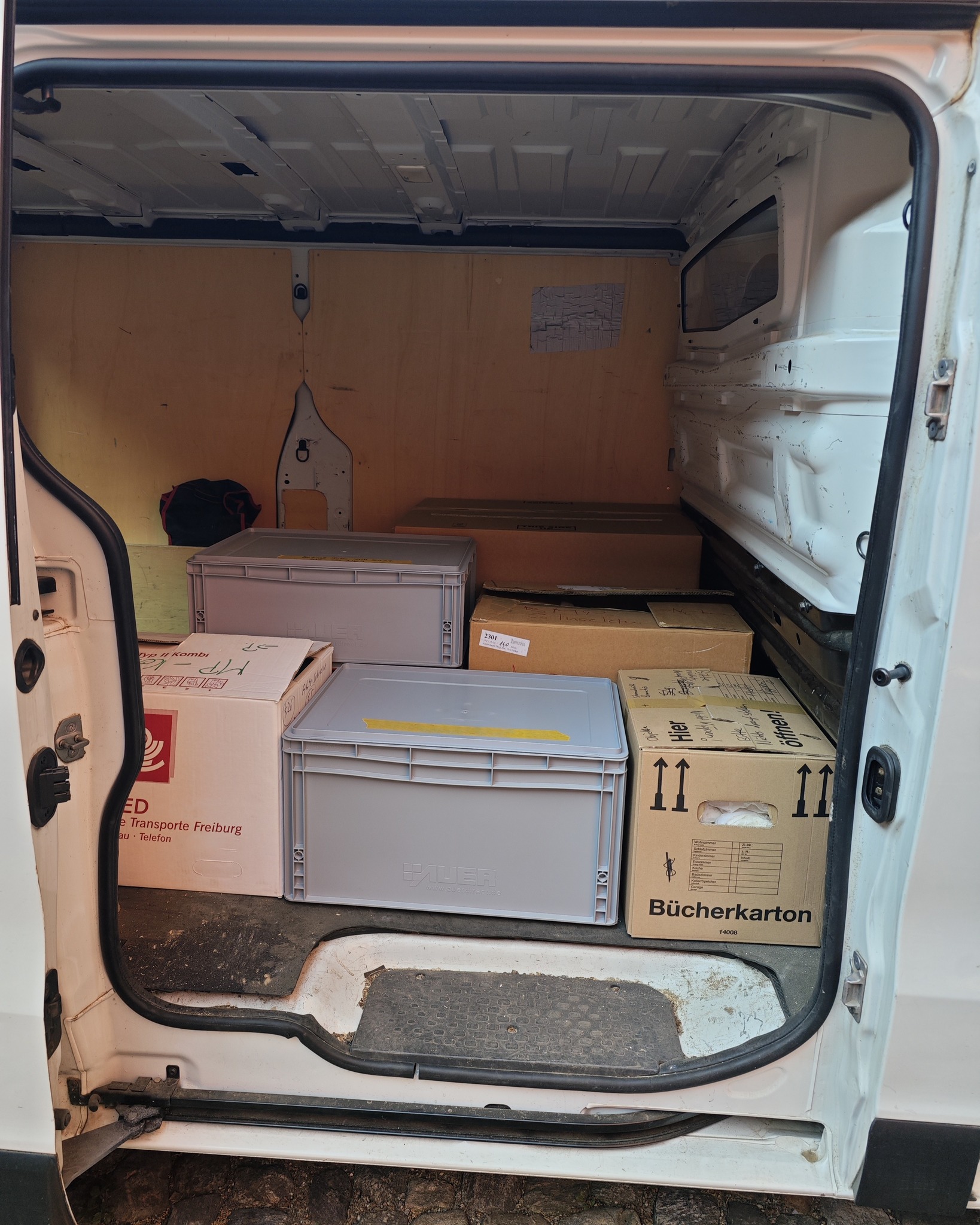 Geöffnete Heckklappe eines weißen Transporters, gefüllt mit gestapelten Pappkartons und grauen Kunststoffkisten. Ein Karton im Vordergrund trägt die Aufschrift „Bücherkarton“, ein weiterer links zeigt den Text „Transporte Freiburg“. (KI-Alternativtext)
