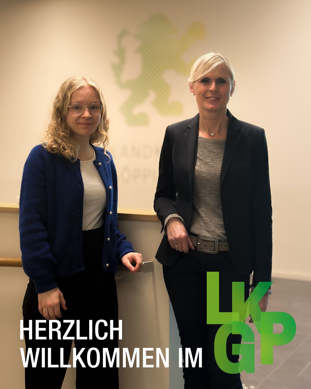 Dieser Alternativtext wurde KI-generiert: Zwei Personen stehen nebeneinander vor einer Wand mit Text. Das Bild ist farbig und im Vordergrund befindet sich ein grünes Logo.