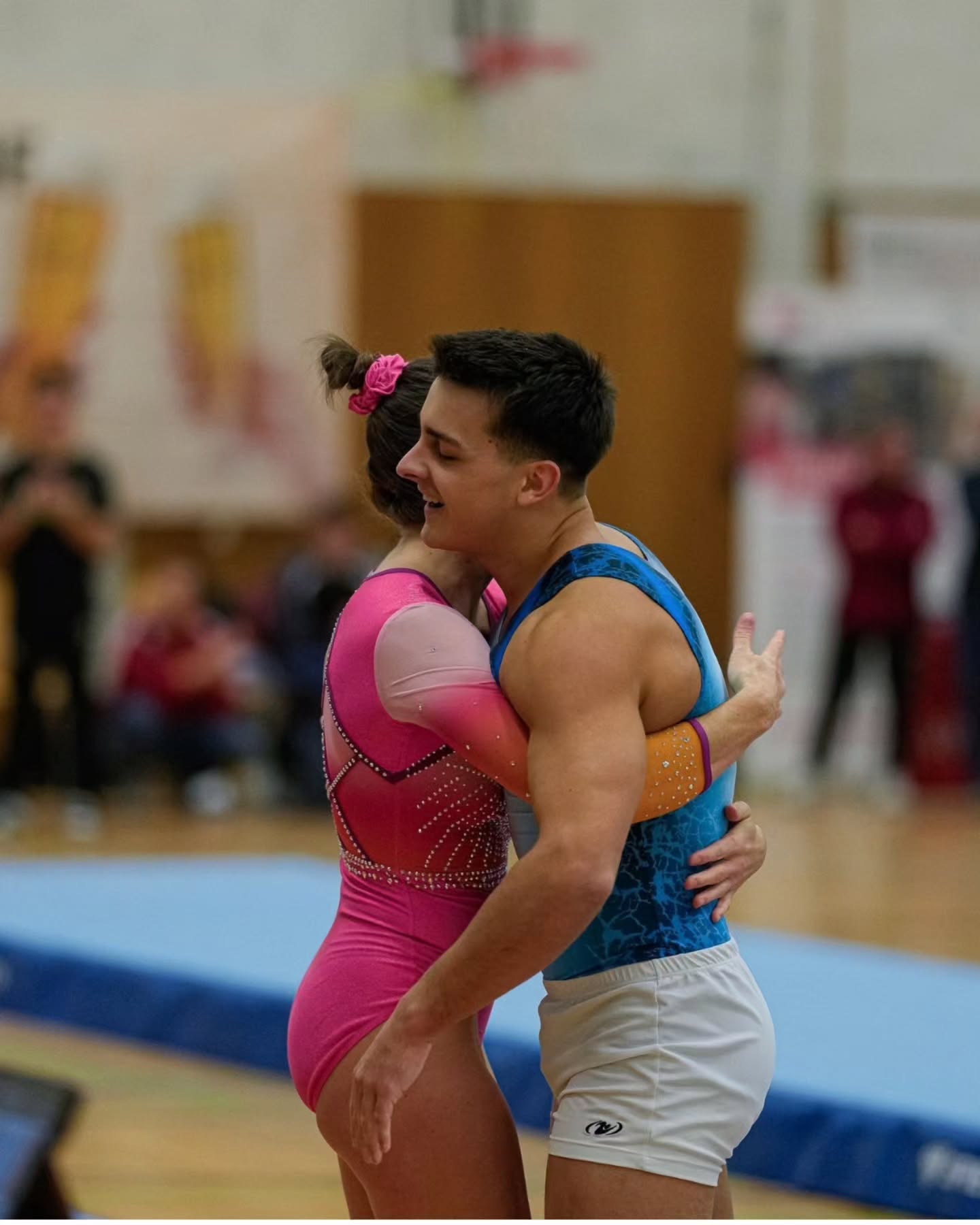 Dieser Alternativtext wurde KI-generiert: Zwei Personen in Sportkleidung umarmen sich vor einem unscharfen Hintergrund einer Sporthalle. Das Bild ist farbig und zeigt eine innige Geste.