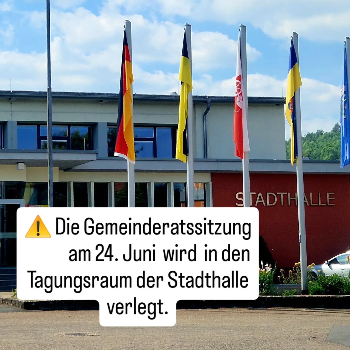 Fassade eines Gebäudes mit der Aufschrift STADTHALLE und mehreren Fahnen. Ein weißer Textkasten mit gelbem Warndreieck informiert: Die Gemeinderatssitzung am 24. Juni wird in den Tagungsraum der Stadthalle verlegt. (KI-Alternativtext)