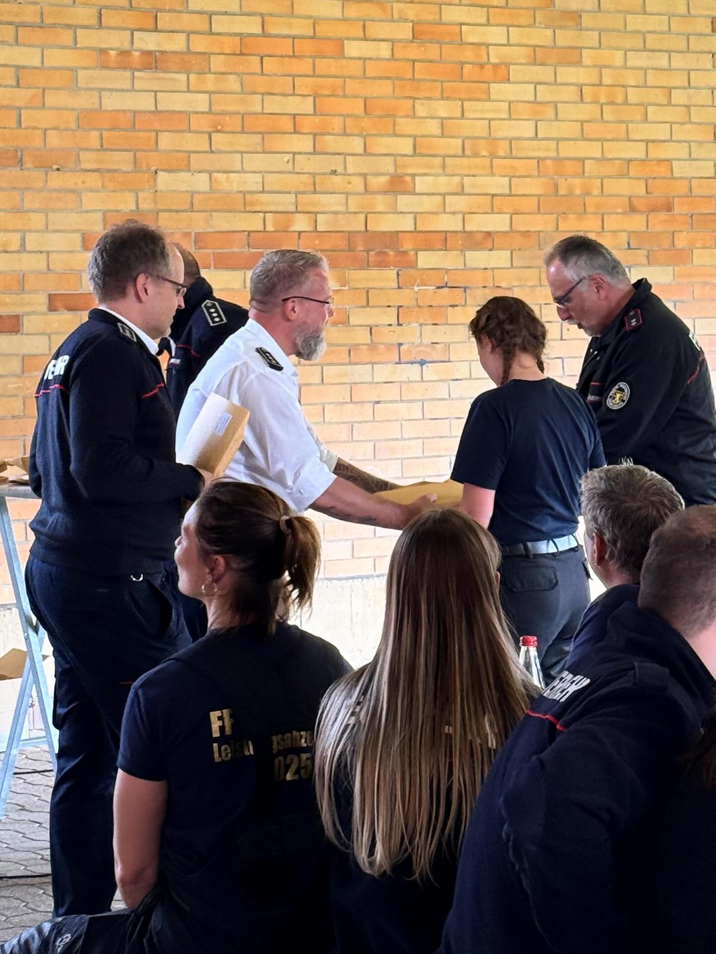 Dieser Alternativtext wurde KI-generiert: Mehrere Personen in Uniformen stehen in einem Raum mit einer Ziegelwand zusammen. Einige halten Gegenstände in den Händen und scheinen miteinander zu kommunizieren.
