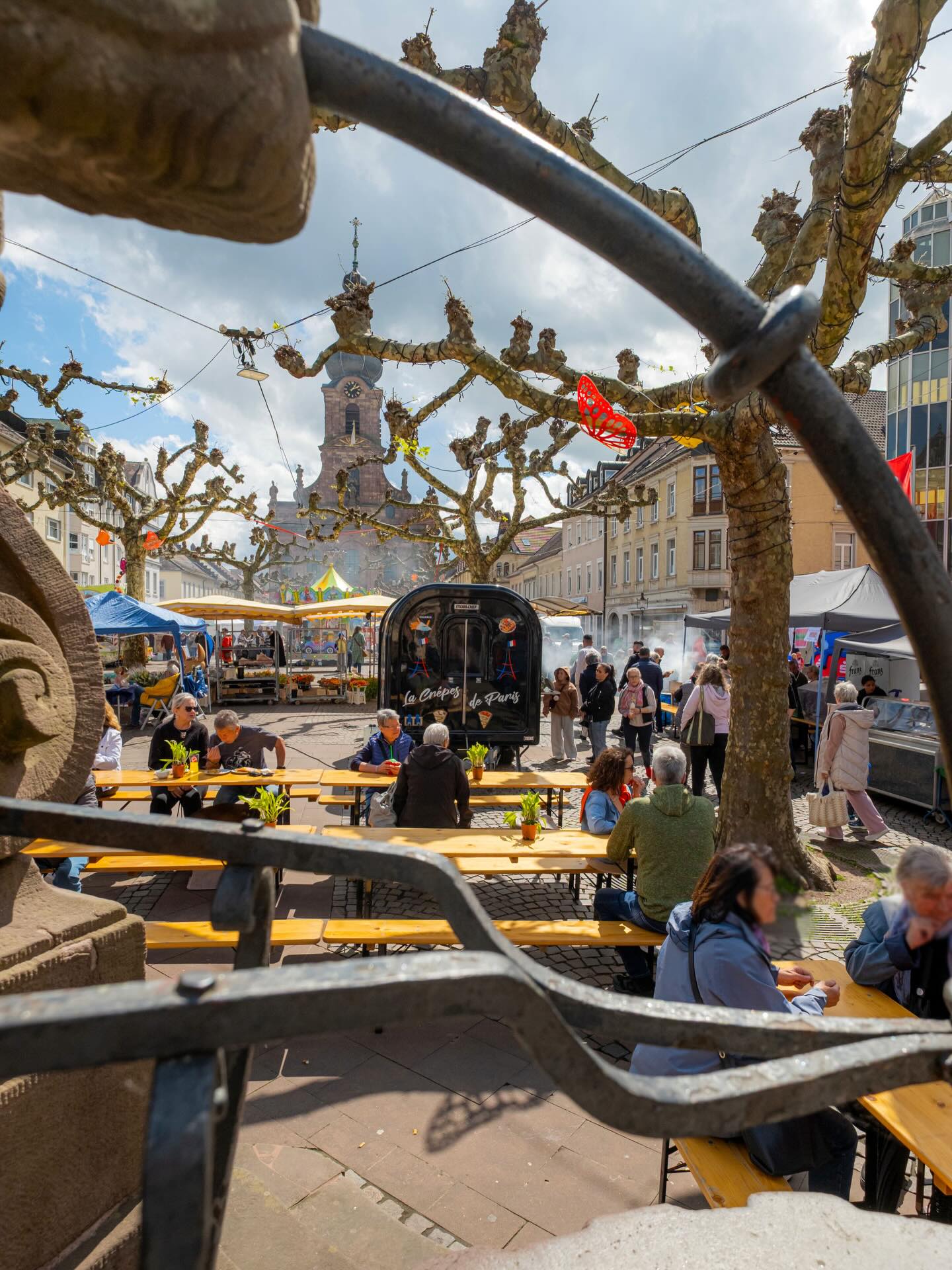 Dieser Alternativtext wurde KI-generiert: Ein Blick durch ein schmiedeeisernes Geländer auf eine belebte Stadtlandschaft mit einem Turm im Hintergrund. Es gibt Essensstände und Menschen an Tischen im Freien.
