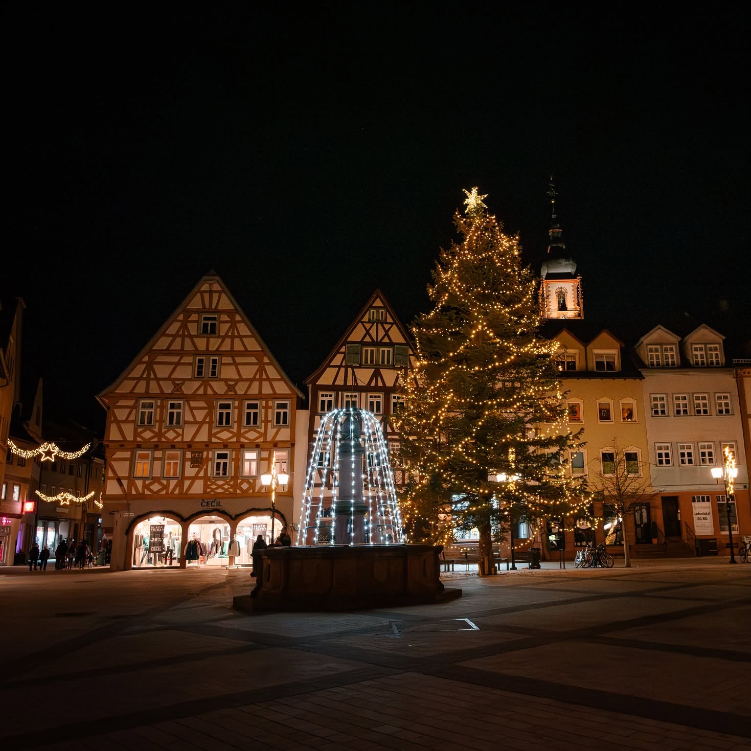 Dieser Alternativtext wurde KI-generiert: Eine nächtliche Aufnahme zeigt einen Platz mit festlicher Beleuchtung, darunter ein beleuchteter Weihnachtsbaum und ein Brunnen. Im Hintergrund sind Gebäude und ein Kirchturm zu sehen.
