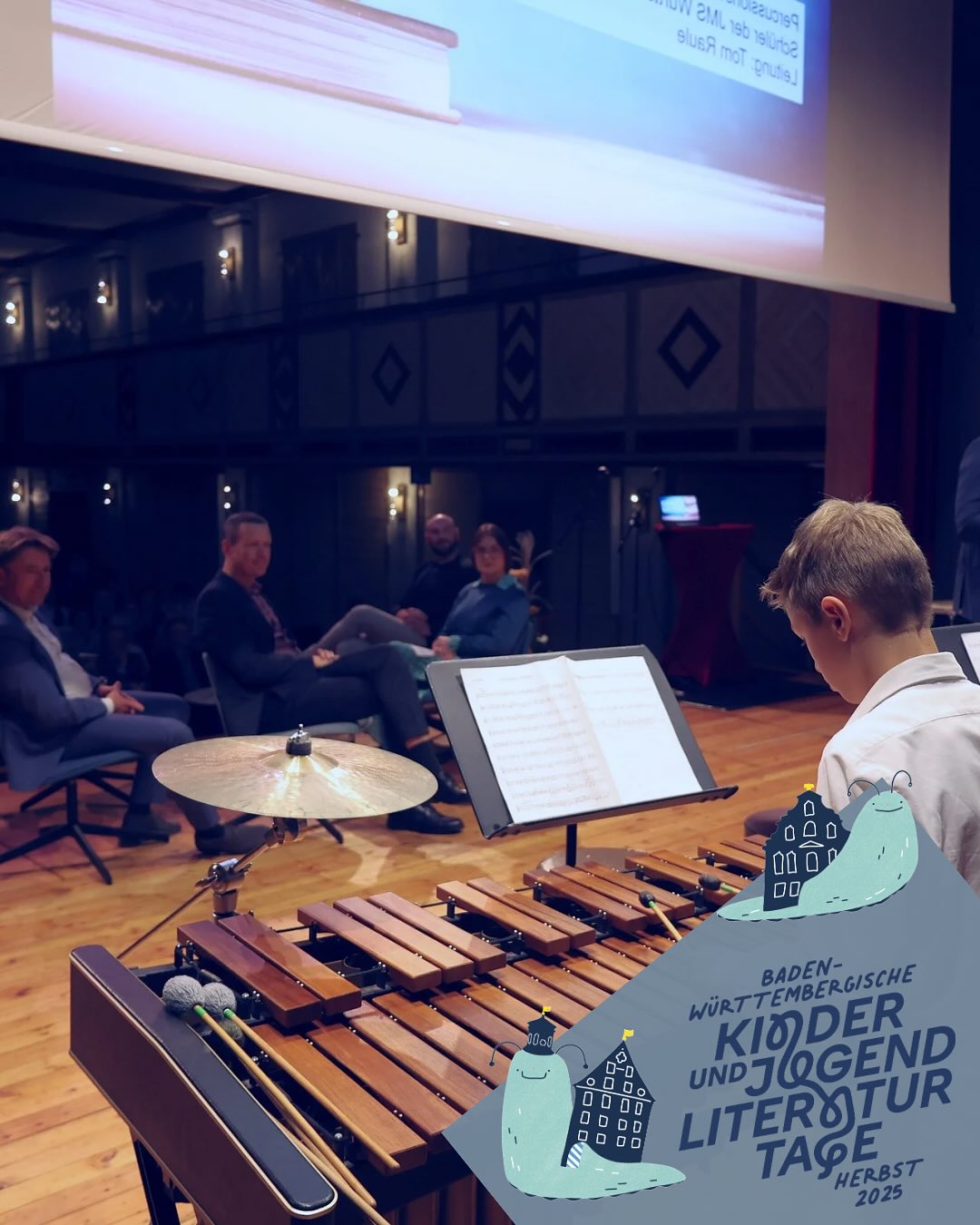 Dieser Alternativtext wurde KI-generiert: Ein Innenraum zeigt eine Bühne mit Musikern und Notenständern. Im Vordergrund befindet sich ein Xylophon und ein Werbeplakat für eine Veranstaltung.