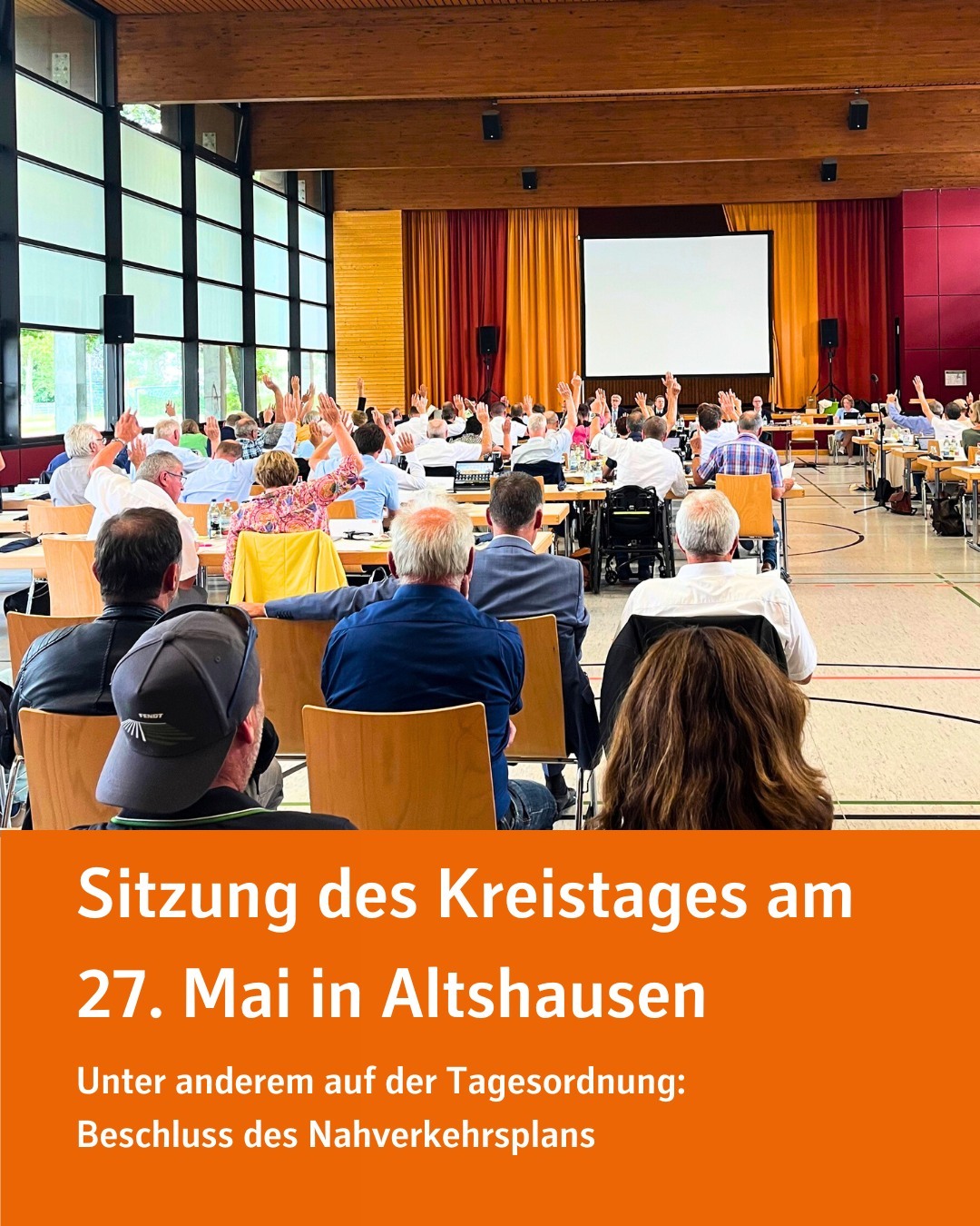 Foto von einer Versammlung in einem großen Raum. Viele Menschen sitzen an Tischen und haben ihre Hände gehoben.