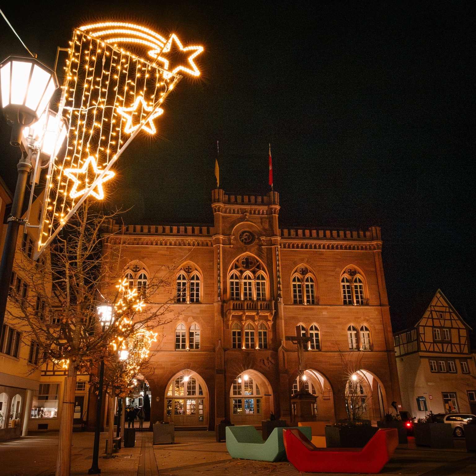 Dieser Alternativtext wurde KI-generiert: Ein nächtliches Stadtbild mit beleuchteten Sternen und einem historischen Gebäude im Hintergrund. Die Szene ist warm durch das Licht der Lichterketten und Laternen erleuchtet.