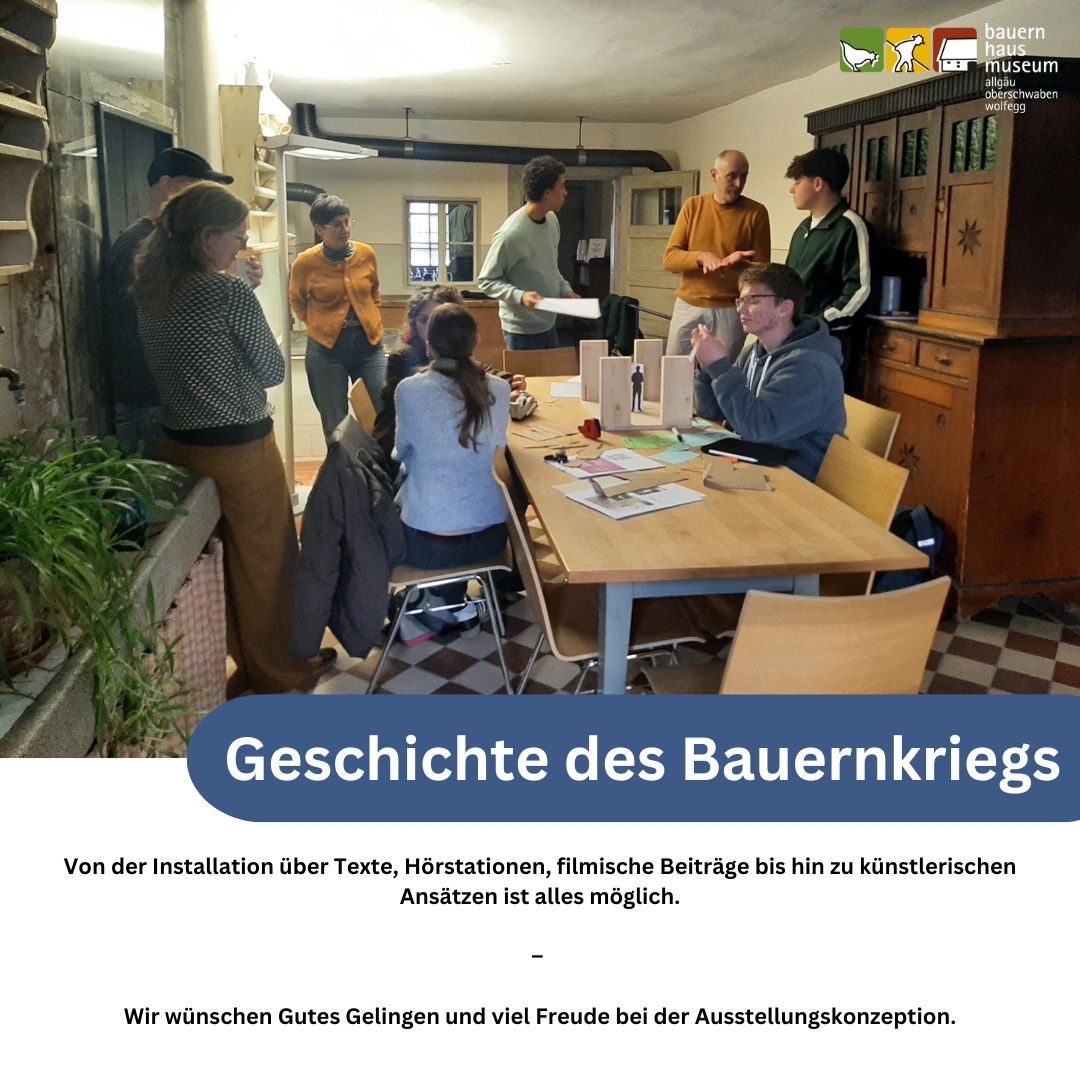 Dieser Alternativtext wurde KI-generiert: Eine Gruppe von Menschen steht und sitzt in einem Innenraum mit einer offenen Tür. Im Vordergrund befindet sich ein Tisch mit Papier, über dem ein Beschriftungsfeld zu sehen ist.