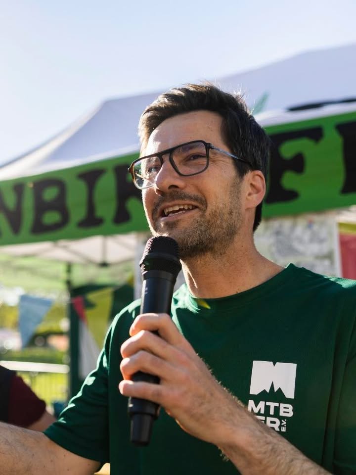 Dieser Alternativtext wurde KI-generiert: Eine Person mit Brille spricht in ein Mikrofon. Im Hintergrund ist ein grüner Banner und ein Zelt zu sehen.