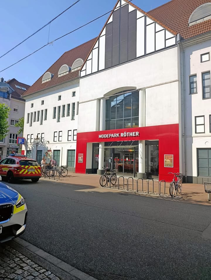 Ein weißes Gebäude mit rotem Fassadenbereich und der Aufschrift "MODEPARK RÖTHER" steht an einer Straße. Vor dem Eingang stehen Fahrradständer mit mehreren Fahrrädern. Links im Bild sind ein teilweise sichtbarer Polizeiwagen und ein Rettungsfahrzeug zu sehen, während zwei Personen auf dem Gehweg spazieren. (KI-Alternativtext)
