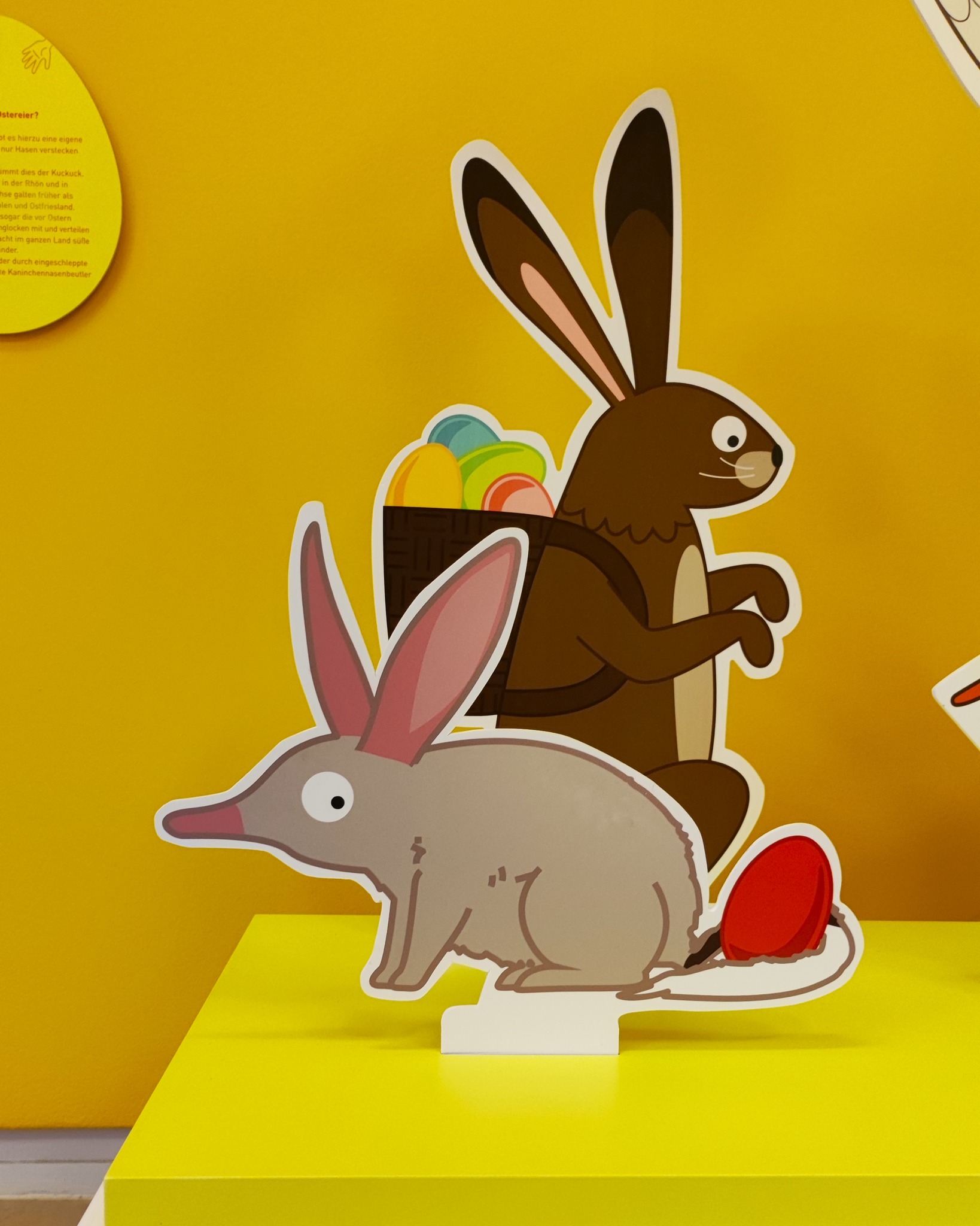 Dieser Alternativtext wurde KI-generiert: Ein helles, farbenfrohes Bild zeigt stilisierte Hasenfiguren aus Pappe und einen Korb mit bunten Eiern. Die Kulisse ist ein gelber Hintergrund mit Text.