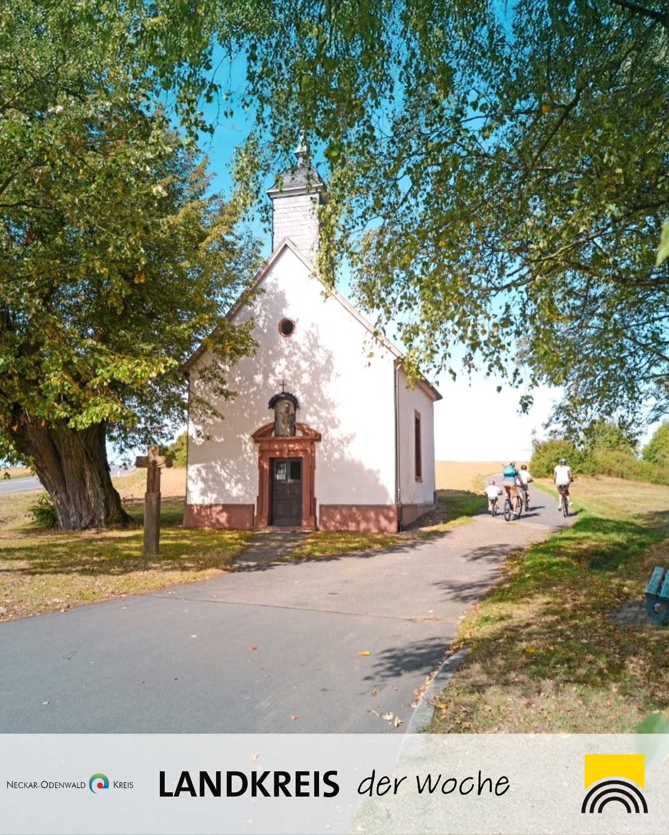 Eine kleine Kapelle steht neben einem Weg. Mehrere Personen fahren mit Fahrrädern auf dem Weg entlang.