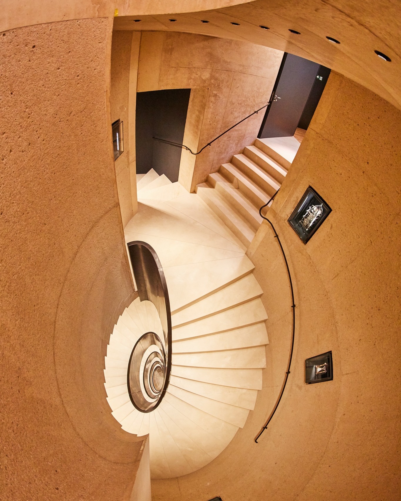 Dieser Alternativtext wurde KI-generiert: Ein spiralförmiges Treppenhaus mit hellen Stufen und einem warmen, texturierten Interieur ist von oben abgebildet. Die Architektur zeichnet sich durch geschwungene Formen und eine offene Gestaltung aus.