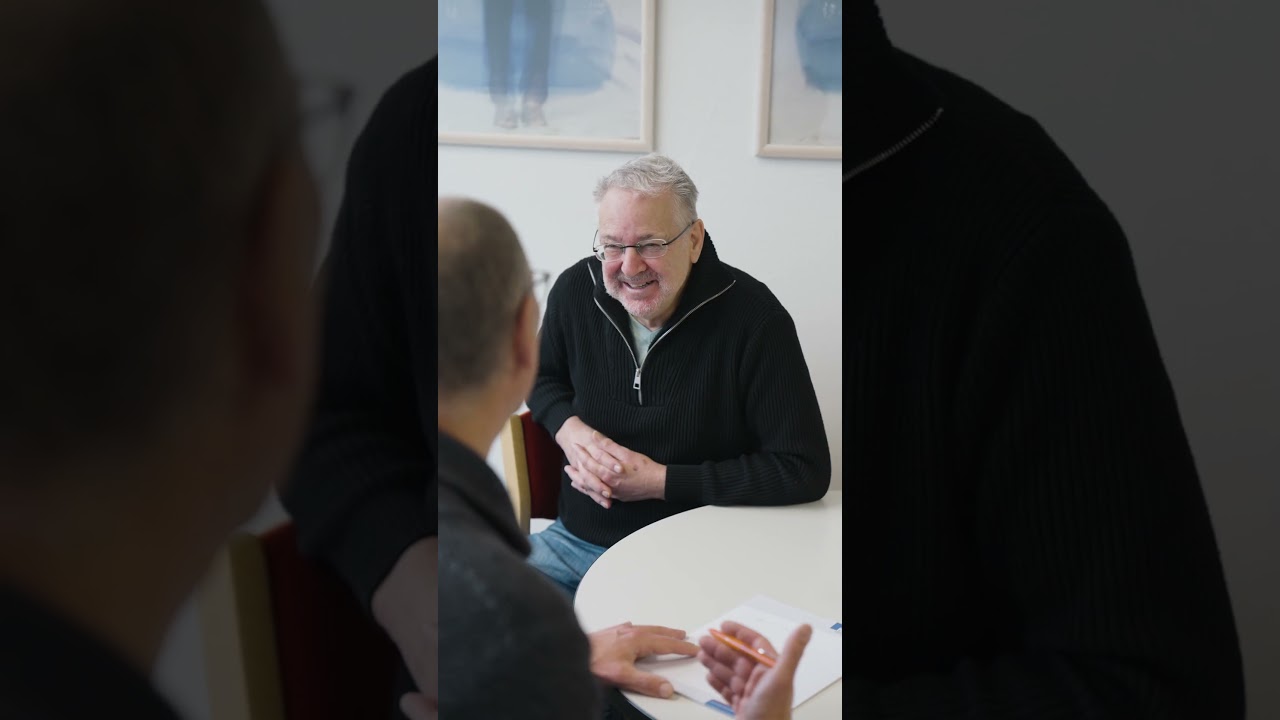 Dieser Alternativtext wurde KI-generiert: Eine Person mit grauen Haaren lächelt und spricht mit zwei weiteren Personen, die teilweise im Bild zu sehen sind. Ein Tisch mit Papieren und einem Stift befindet sich im Fokus.
