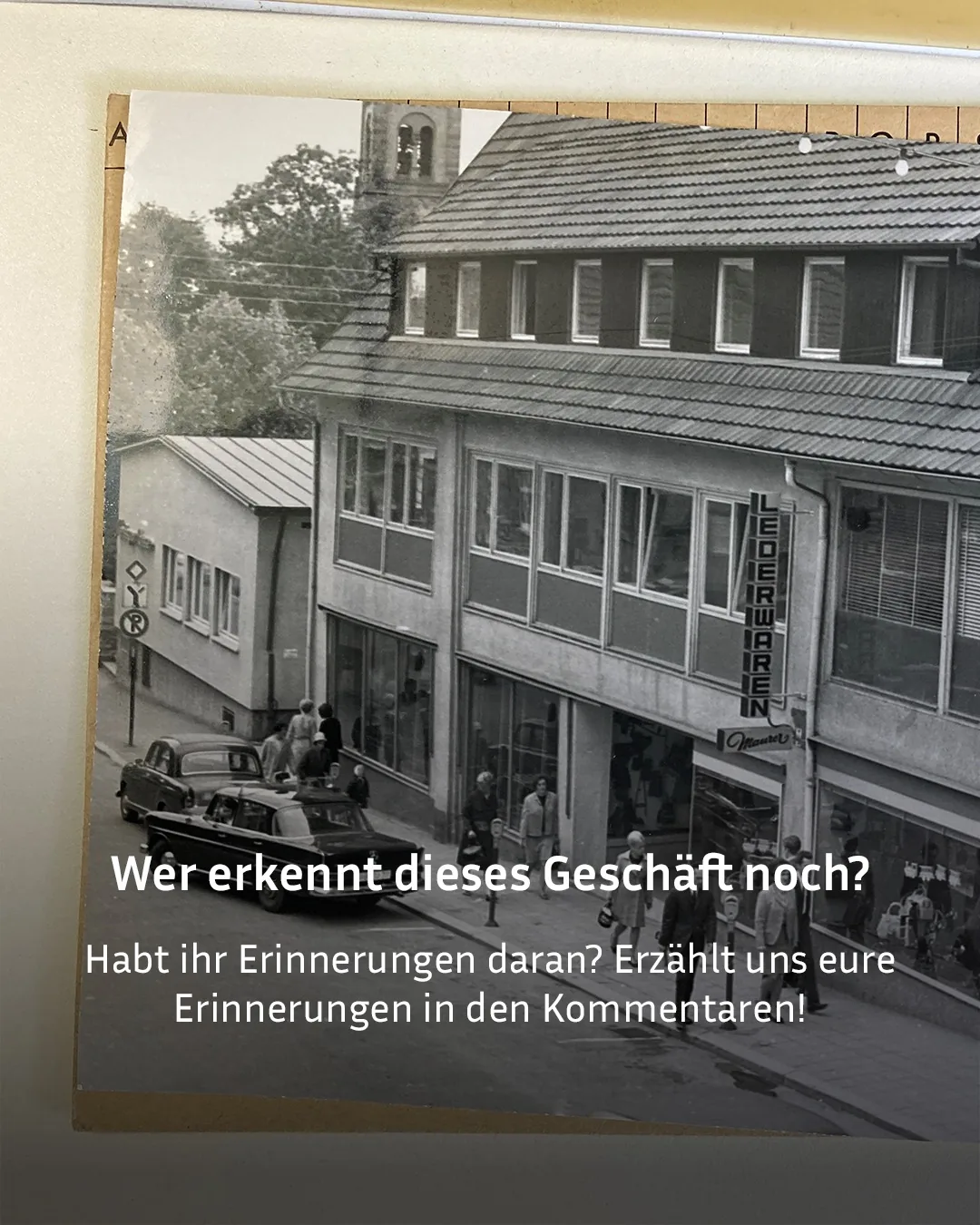 Schwarzweißfoto einer Straße mit einem Gebäude, dessen Fassade einen senkrechten Schriftzug „LEDERWAREN“ und ein Schild „Maurer“ zeigt. Mehrere Autos der alten Bauzeit stehen auf der Straße, Menschen gehen auf dem Gehweg oder stehen an den Fahrzeugen. Im Hintergrund sind Bäume und ein Kirchturm zu sehen. Der eingeblendete Text lautet: „Wer erkennt dieses Geschäft noch? Habt ihr Erinnerungen daran? Erzählt uns eure Erinnerungen in den Kommentaren!" (KI-Alternativtext)