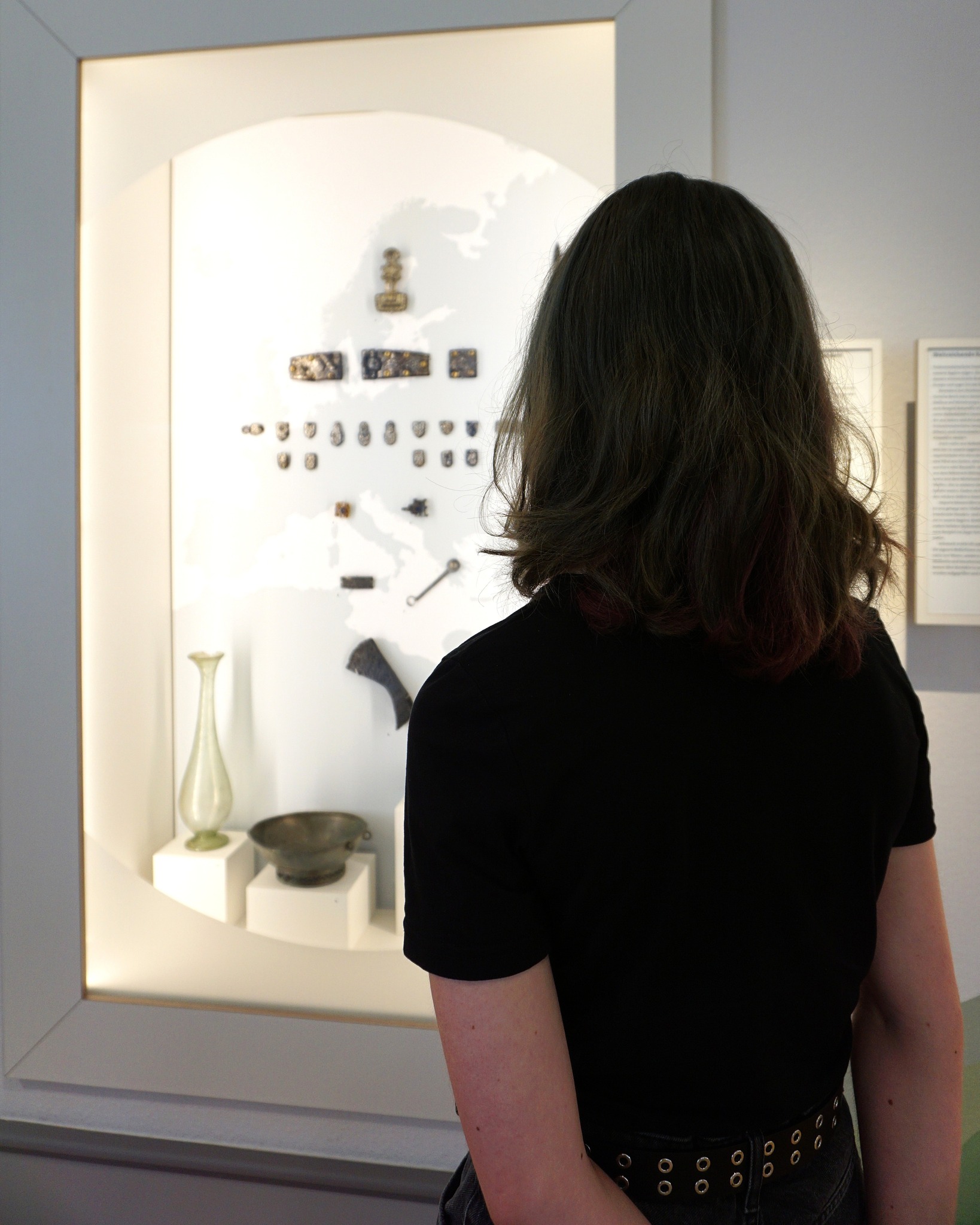 Eine Person mit dunklen Haaren und schwarzem T-Shirt steht vor einer beleuchteten Museumsvitrine. In der Vitrine sind auf weißem Hintergrund verschiedene Gegenstände ausgestellt, darunter Metallplatten, kleine Metallteile, eine Glasvase und eine Schale. Rechts an der Wand sind Texttafeln angebracht. (KI-Alternativtext)