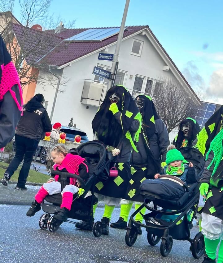 Dieser Alternativtext wurde KI-generiert: Eine Gruppe von Personen in auffälligen, pelzigen Kostümen und leuchtend grünen Elementen befindet sich auf einer Straße vor einem Gebäude. Mehrere Kinder sind in Kinderwagen oder werden gezogen.