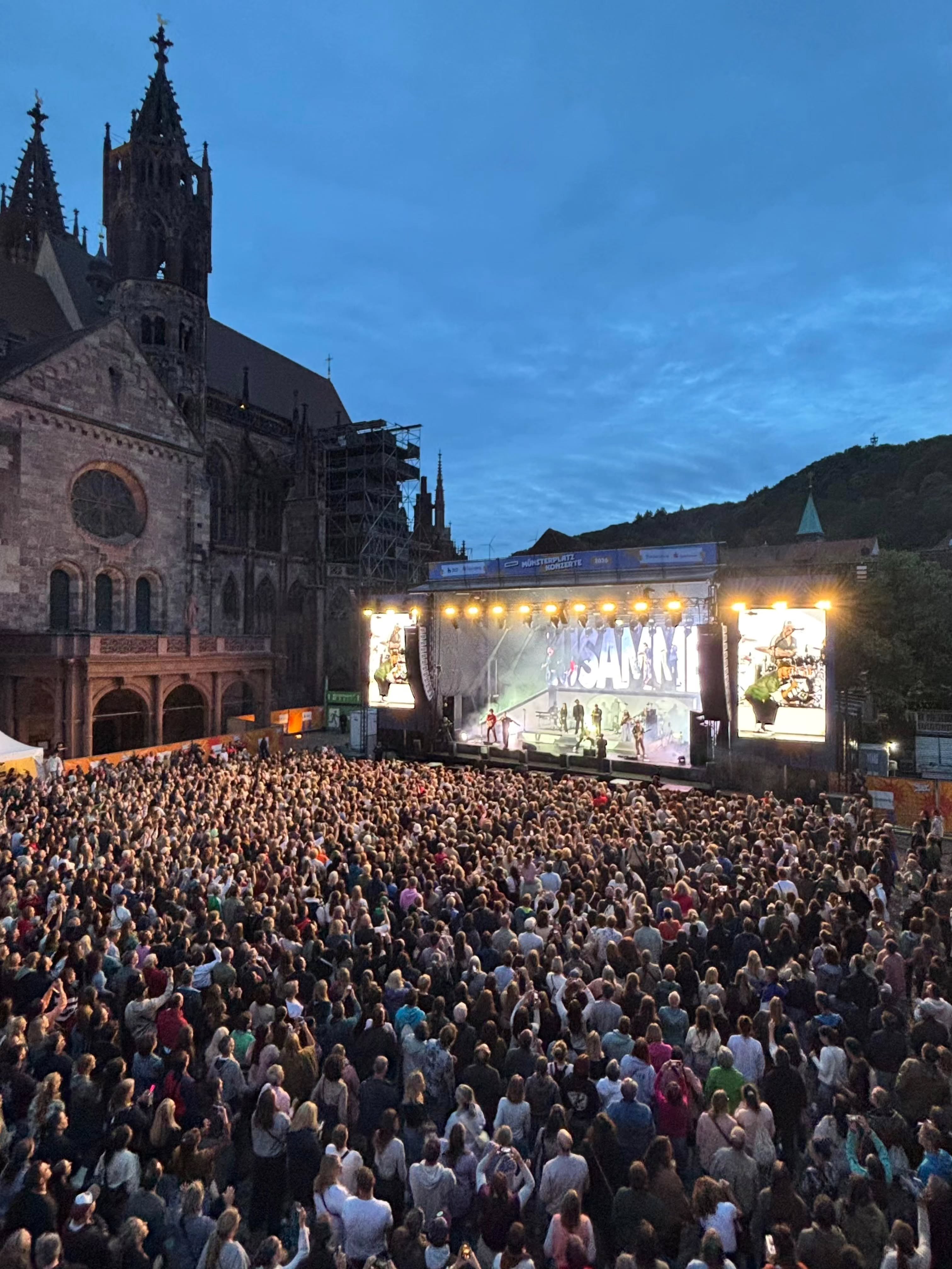 Große Menschenmenge vor einer beleuchteten Bühne mit Musiker:innen und großen Bildschirmen, im Hintergrund eine gotische Kirche bei Dämmerung. Auf der Bühne ist der Schriftzug „SAMI“ zu sehen. (KI-Alternativtext)