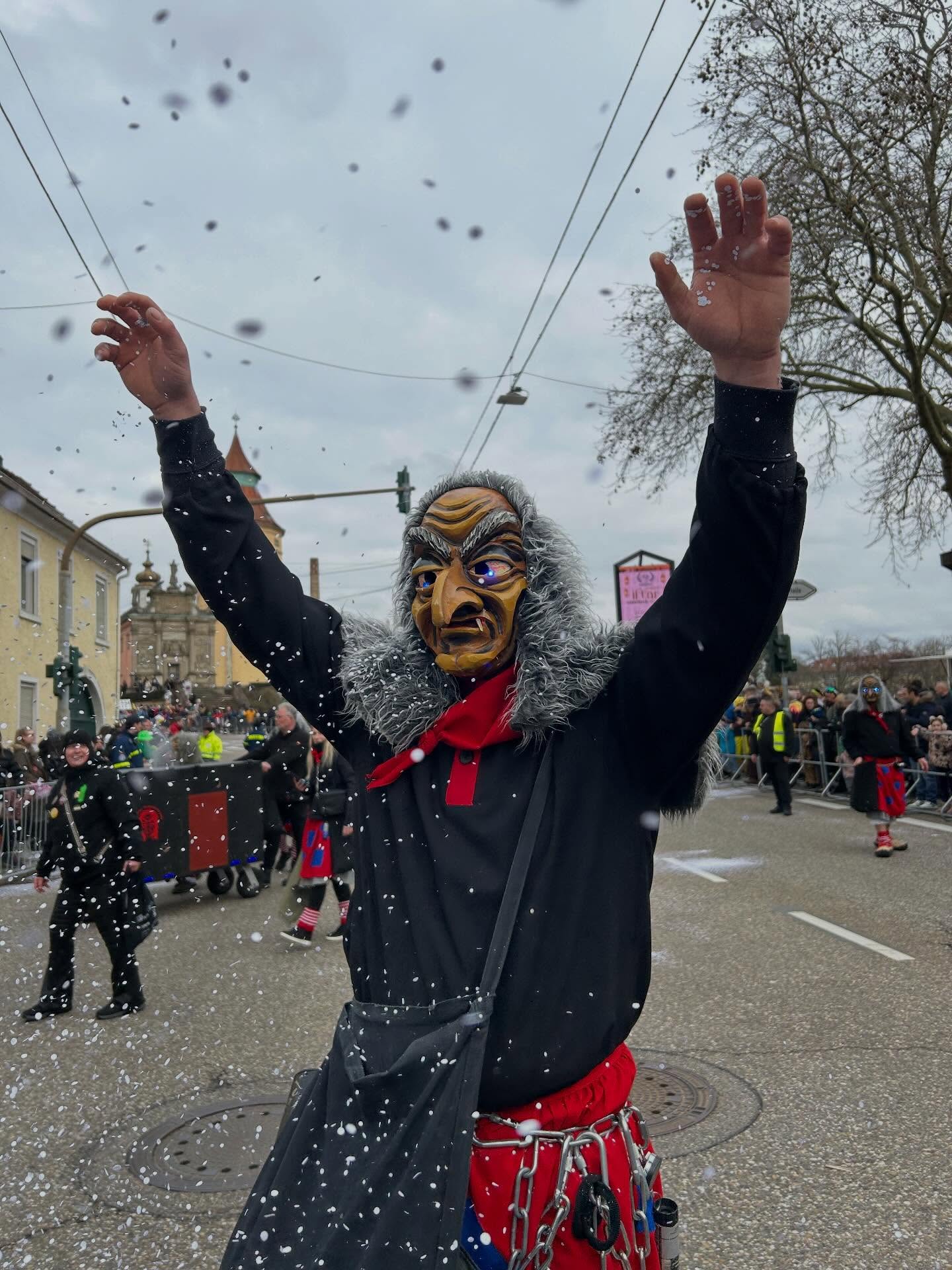 Dieser Alternativtext wurde KI-generiert: Eine Person in einem auffälligen, traditionellen Kostüm mit einer Maske steht im Schnee und hat die Arme erhoben. Im Hintergrund sind weitere Menschen und Gebäude zu sehen.