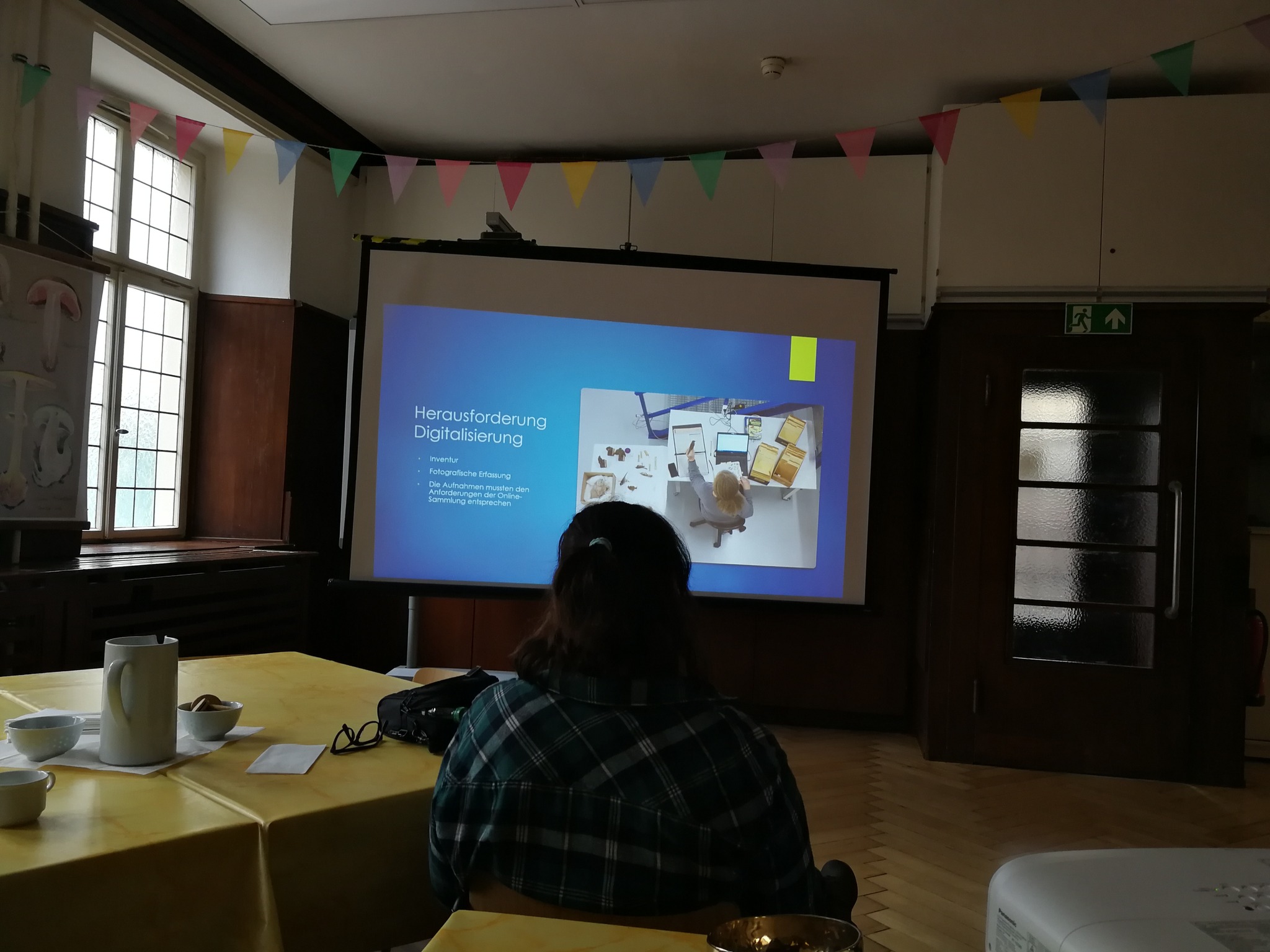 Eine Person sitzt im Rücken vor einer Leinwand mit einer Präsentation über die Herausforderung der Digitalisierung. Auf dem Tisch stehen Getränke und Gebäck.