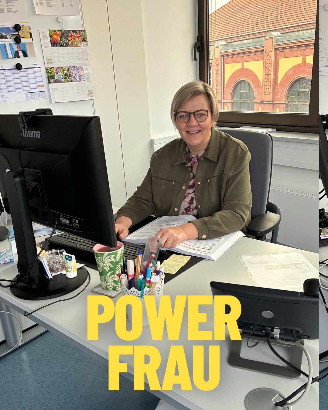 Dieser Alternativtext wurde KI-generiert: Ein Arbeitsplatz mit einem Schreibtisch, einem Computer und verschiedenen Büromaterialien. Im Vordergrund sitzt eine Person an dem Schreibtisch und lächelt.