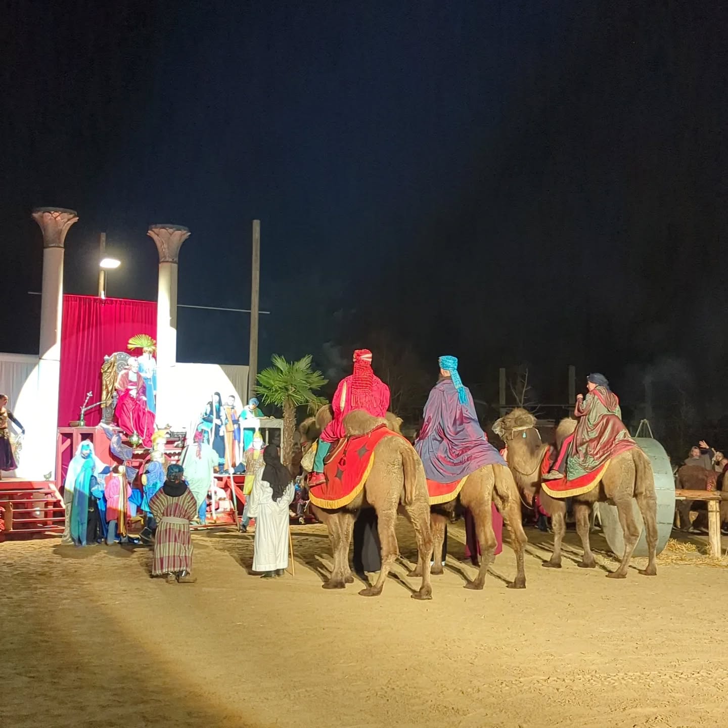 Dieser Alternativtext wurde KI-generiert: Eine nächtliche Szene zeigt mehrere Personen auf Kamelen und zu Fuß vor einer roten Kulisse. Pflanzen und Strukturen sind im Hintergrund zu sehen.