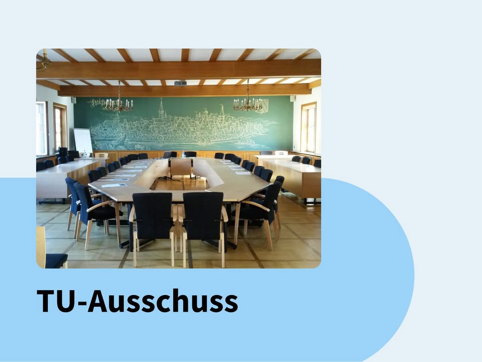 Dieser Alternativtext wurde KI-generiert: Ein Raum mit einem langen Tisch und Stühlen, an dessen hinterer Wand eine große Tafel mit einer städtischen Szene hängt. Der Raum hat eine helle, freundliche Atmosphäre.