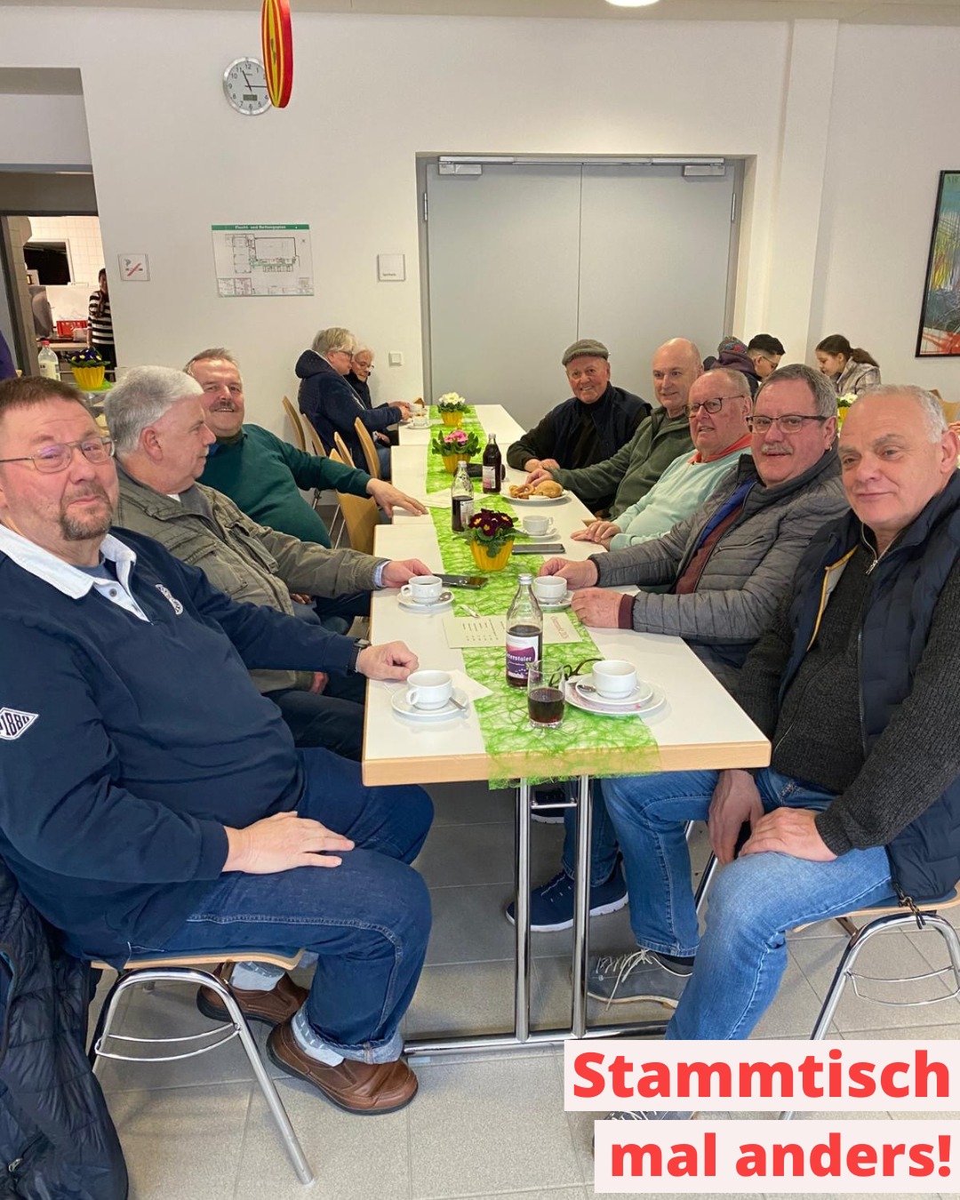 Dieser Alternativtext wurde KI-generiert: Eine Gruppe von Menschen sitzt an einem Tisch mit Getränken und Snacks in einem hellen Raum. Im Hintergrund befindet sich eine Tür und eine Wegweiser-Tafel.