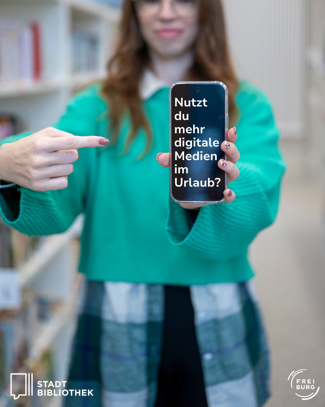 Eine Person hält ein Smartphone hoch und zeigt mit dem Finger auf den Bildschirm, auf dem der Text „Nutzt du mehr digitale Medien im Urlaub?“ steht. Im Hintergrund sind unscharf Bücherregale zu sehen. Unten links ist das Logo der Stadtbibliothek und unten rechts das Logo von Freiburg abgebildet. (KI-Alternativtext)