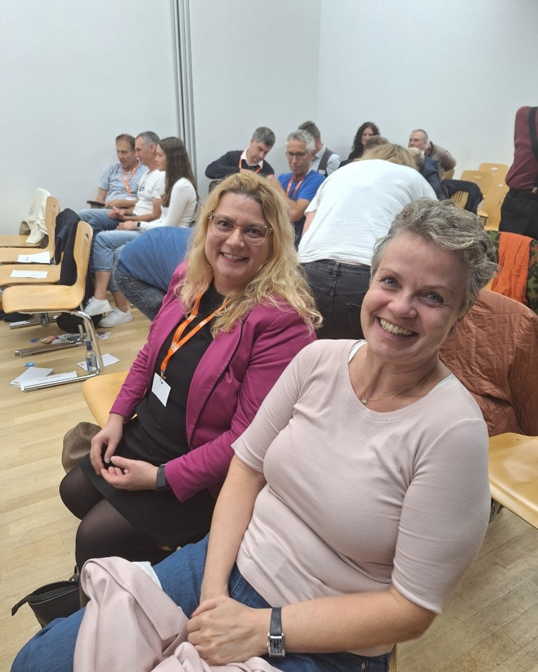 Zwei Frauen (Tina Schädler und Dr. Barbara Segelbacher) sitzen auf Stühlen in Publikumsstuhlreihen und lächeln ins Blickfeld. Im Hintergrund andere sitzende Menschen.
