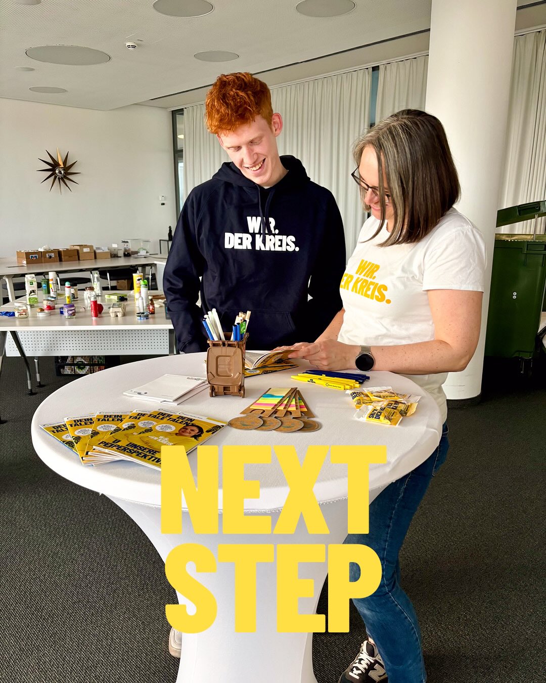 Dieser Alternativtext wurde KI-generiert: Zwei Personen stehen an einem Tisch mit Materialien und vorbereiten etwas. Im Hintergrund ist ein großes Schild mit der Aufschrift “NEXT STEP” zu sehen.