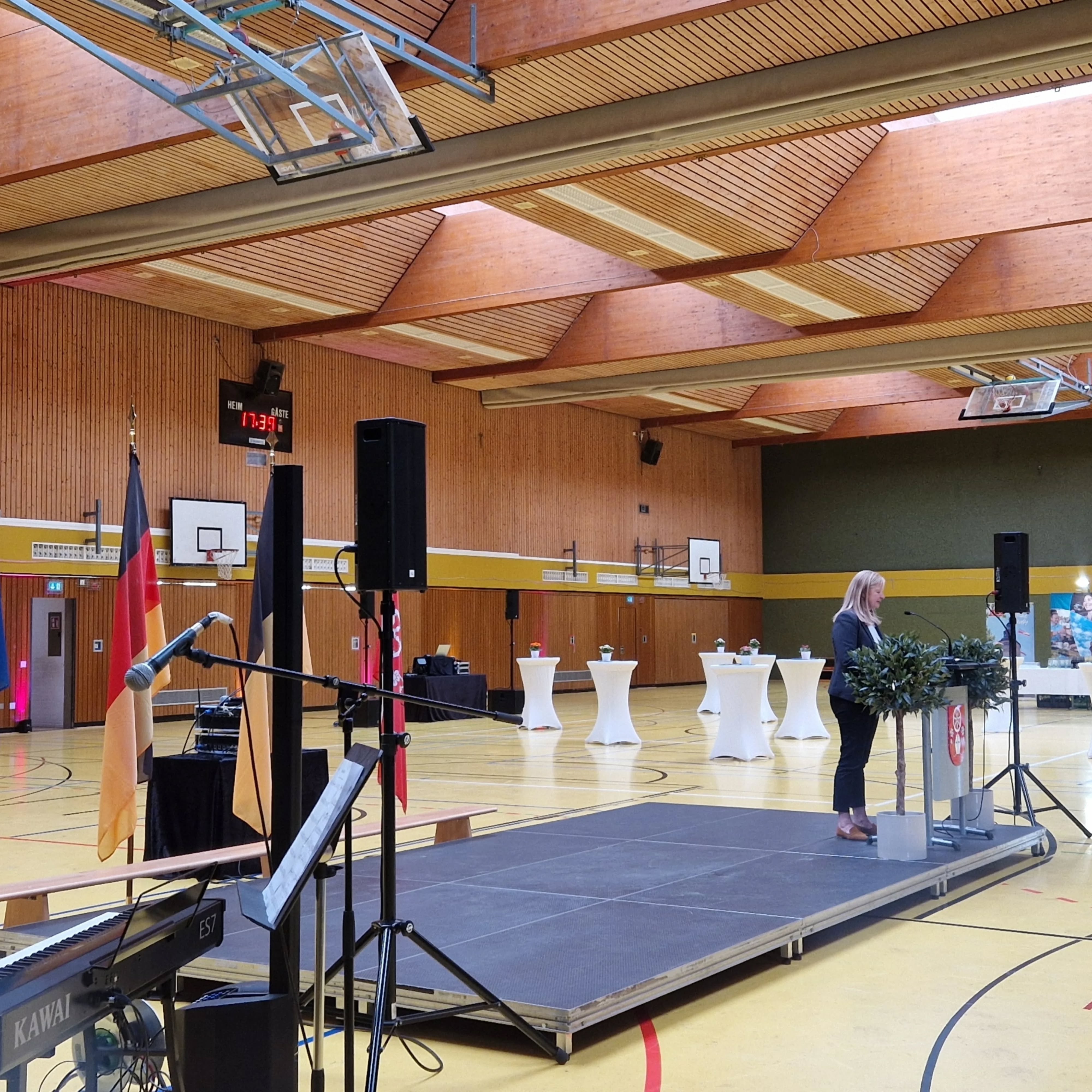 Dieser Alternativtext wurde KI-generiert: Ein helles Rauminnere mit Holzwänden und -decke. Eine Person steht auf einer erhöhten Bühne und spricht in ein Mikrofon, während im Hintergrund eine Sporthalle zu sehen ist.